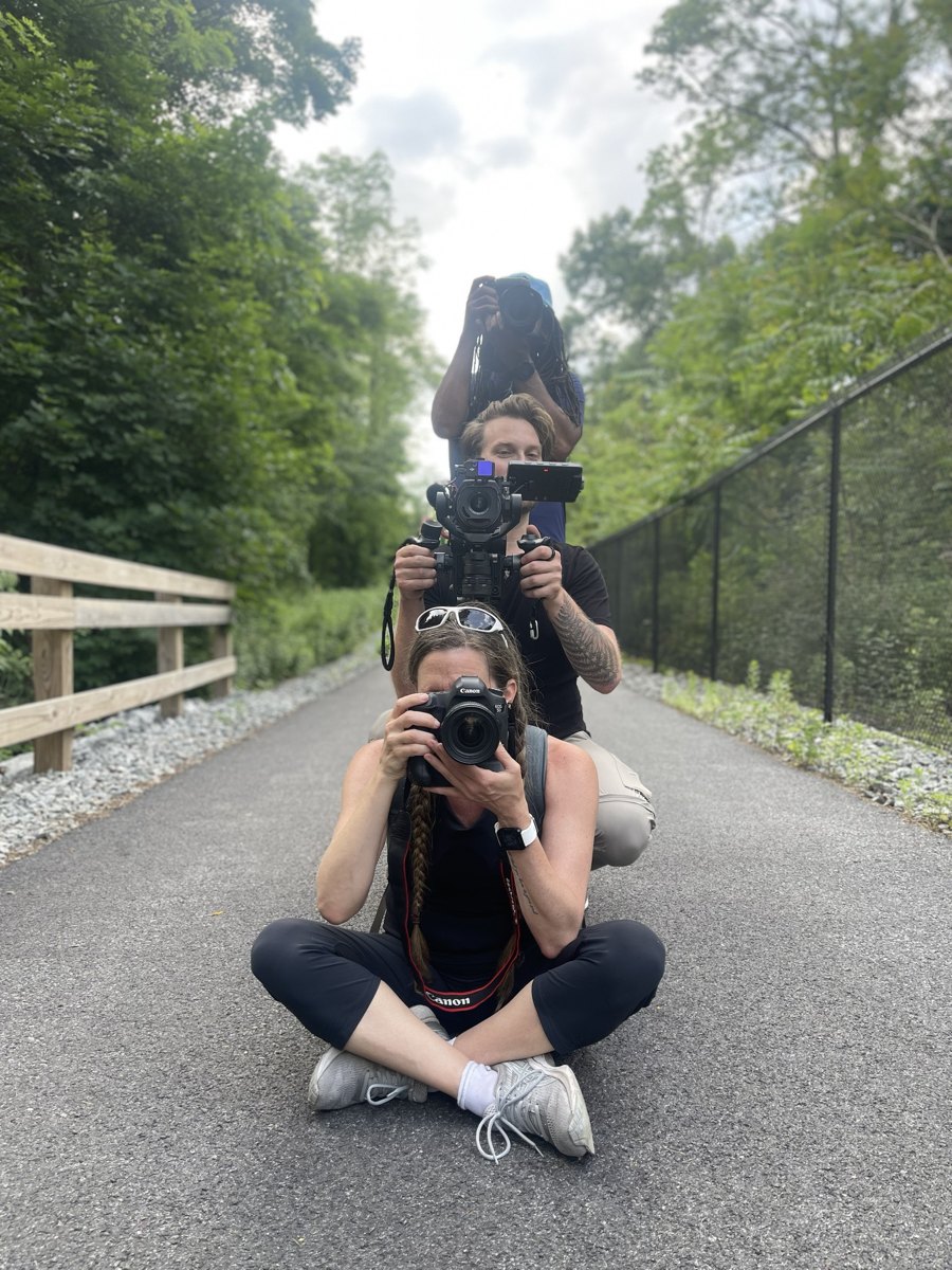 Three people stacked one behind another on a paved trail, each holding a camera toward the lens, Tyler in the middle with a gimbal