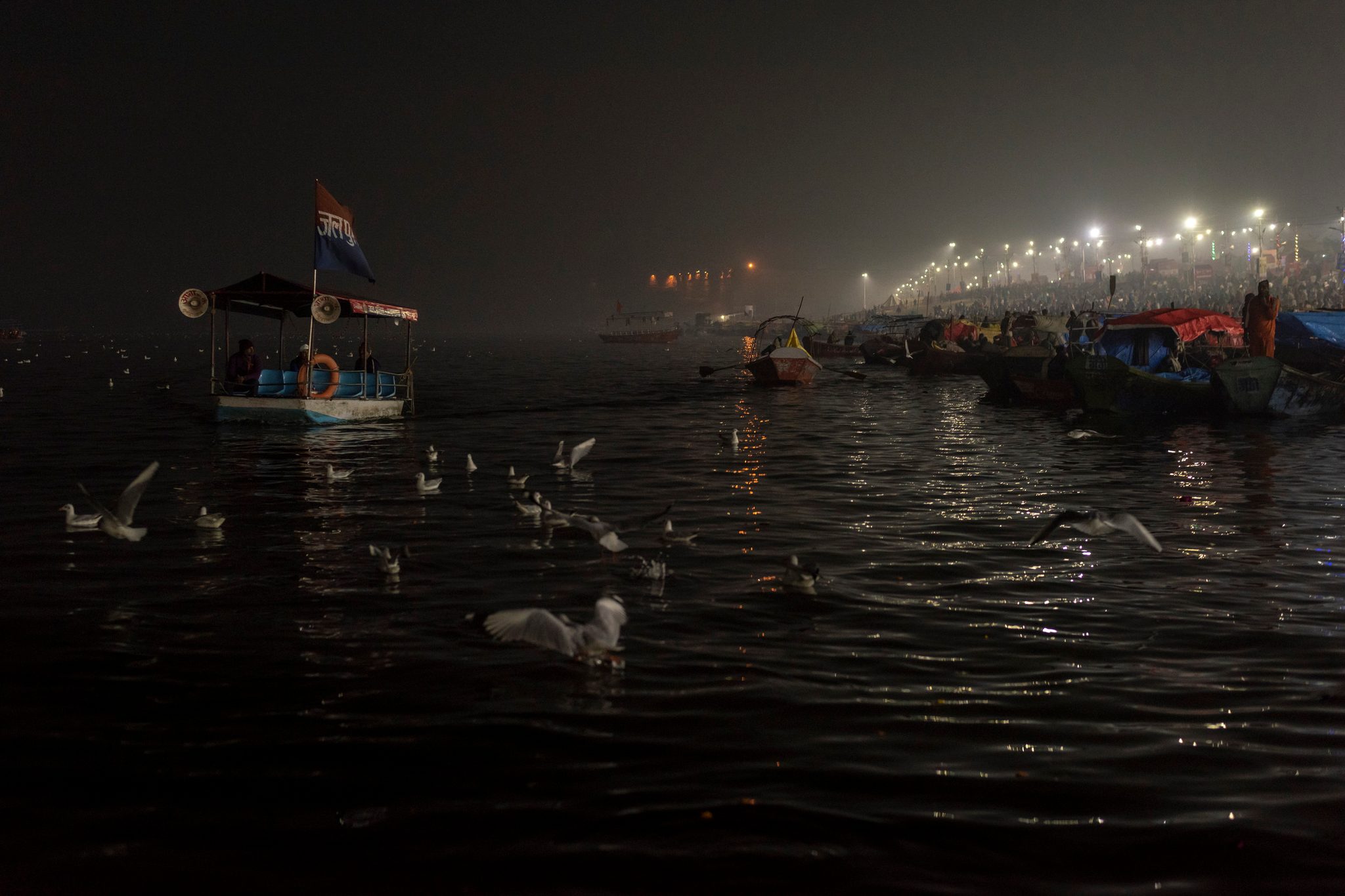 Boats wait on the dark river as gulls settle on the water, the far shore lit end to end.
