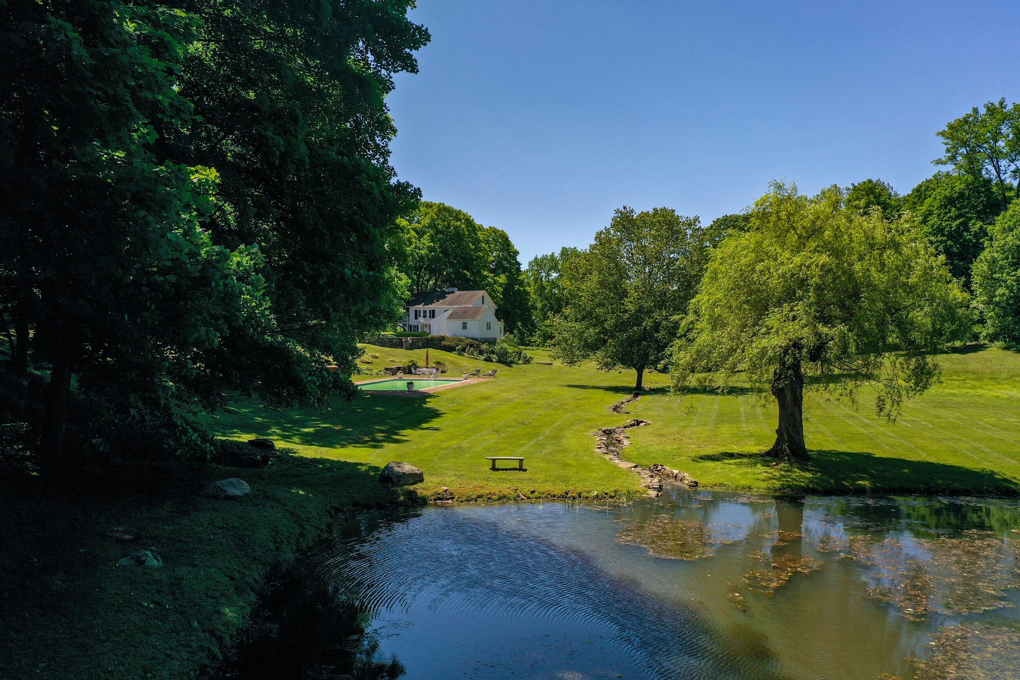 A June morning from above, with a weeping willow leaning over the pond and a mown path along the water.