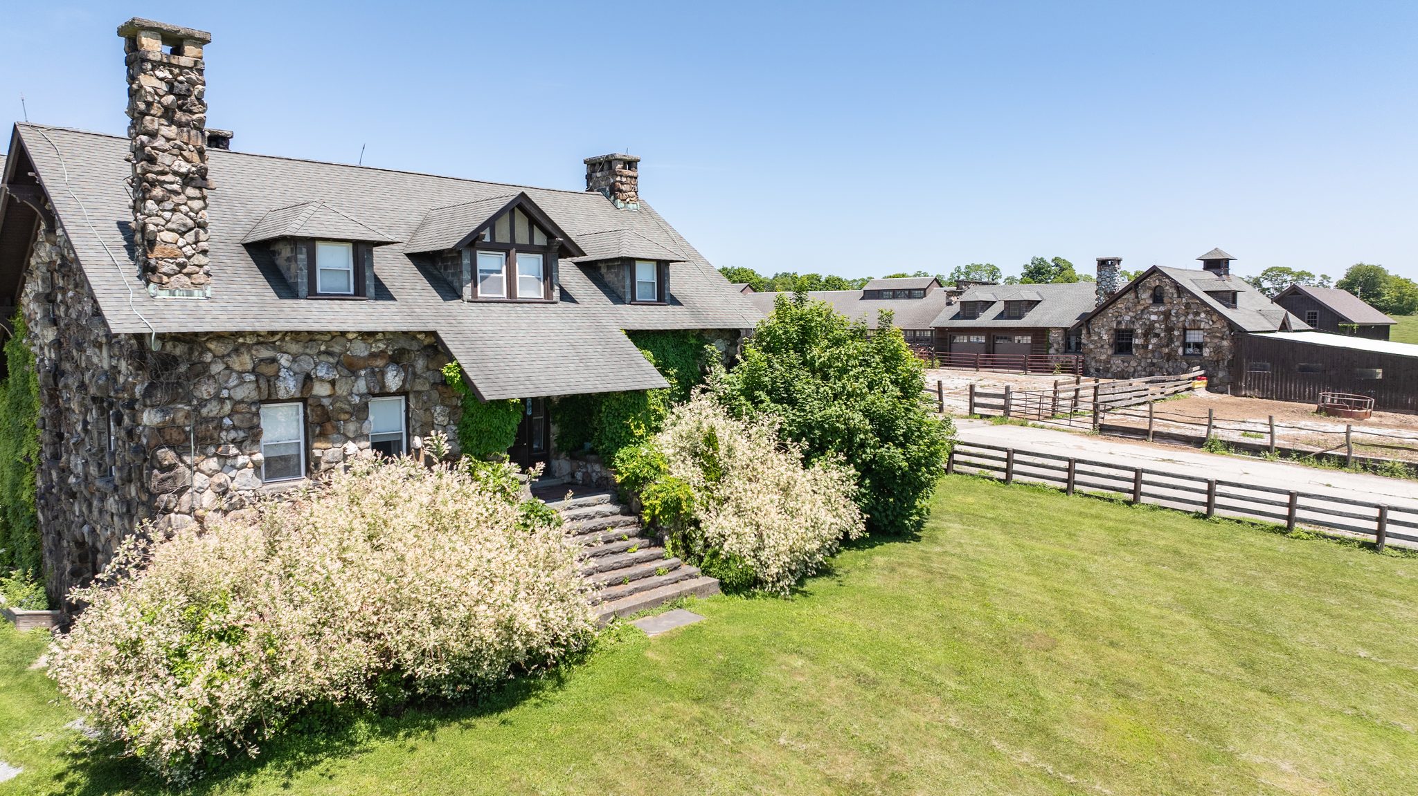 Fieldstone stables with cupolas and rail-fenced paddocks spread out under a clear early June sky, seen from the drone.