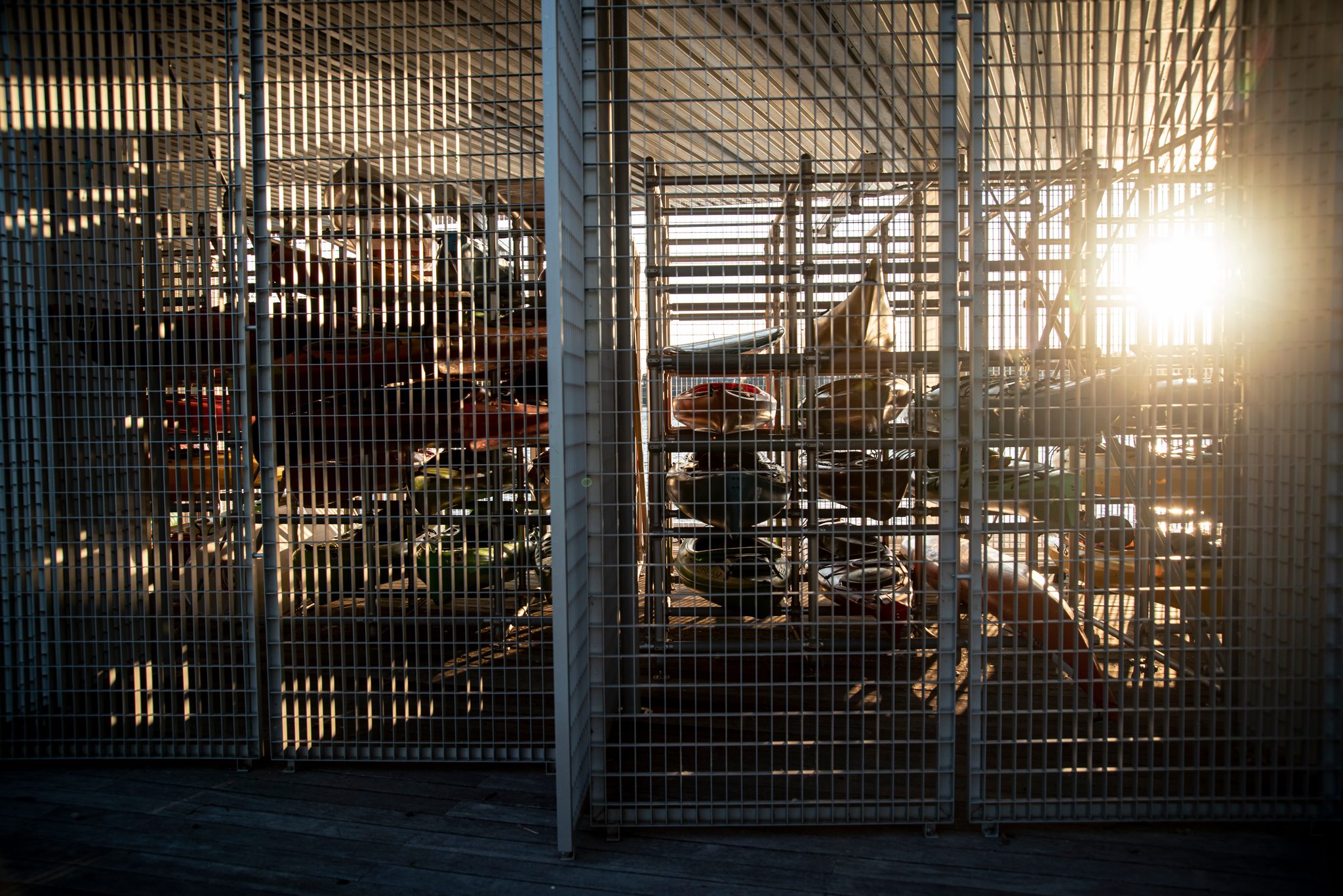 Kayaks rest in their steel racks behind the mesh, the late sun slipping through to find them.