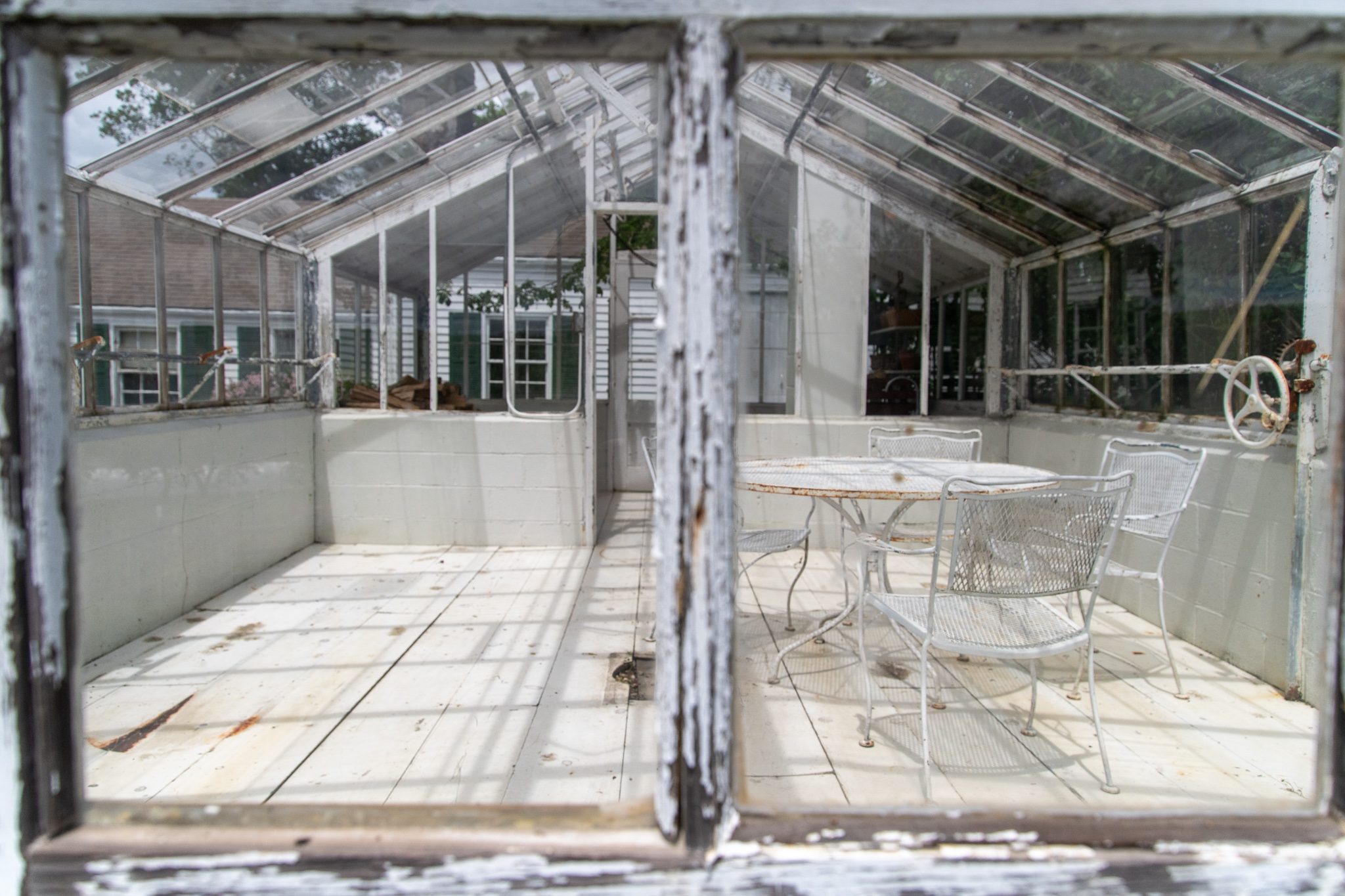 Through a peeling window frame, an old glass greenhouse holds a table and chairs.