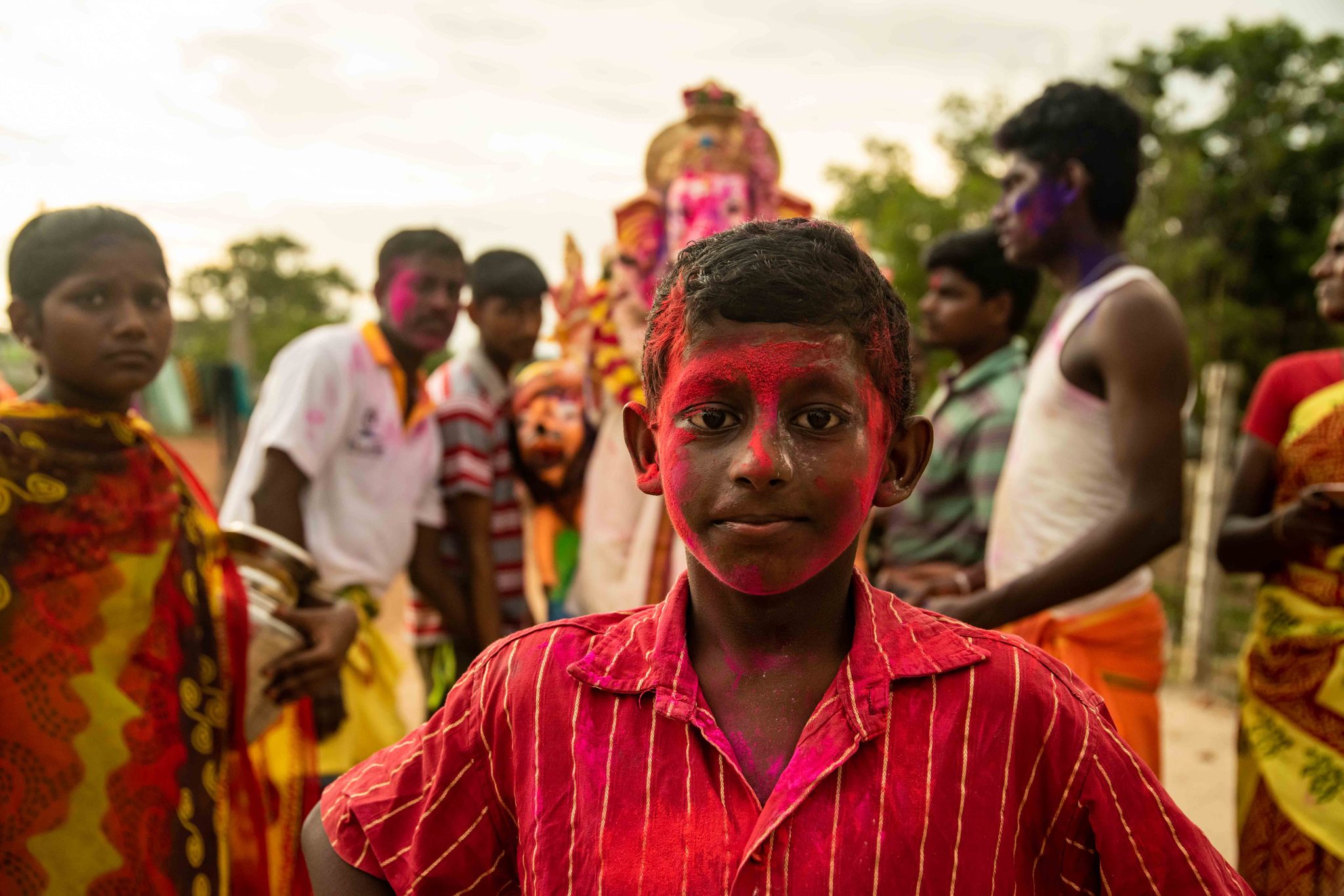 A boy with a face full of pink powder stands before the procession.