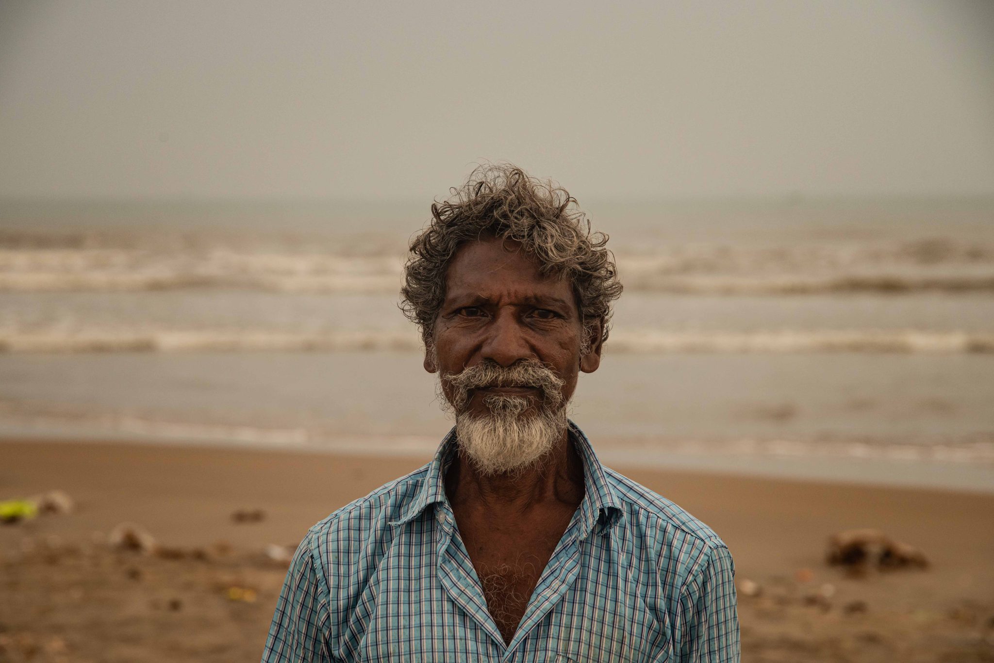 A fisherman with a white beard stands on the beach, the waves behind him.