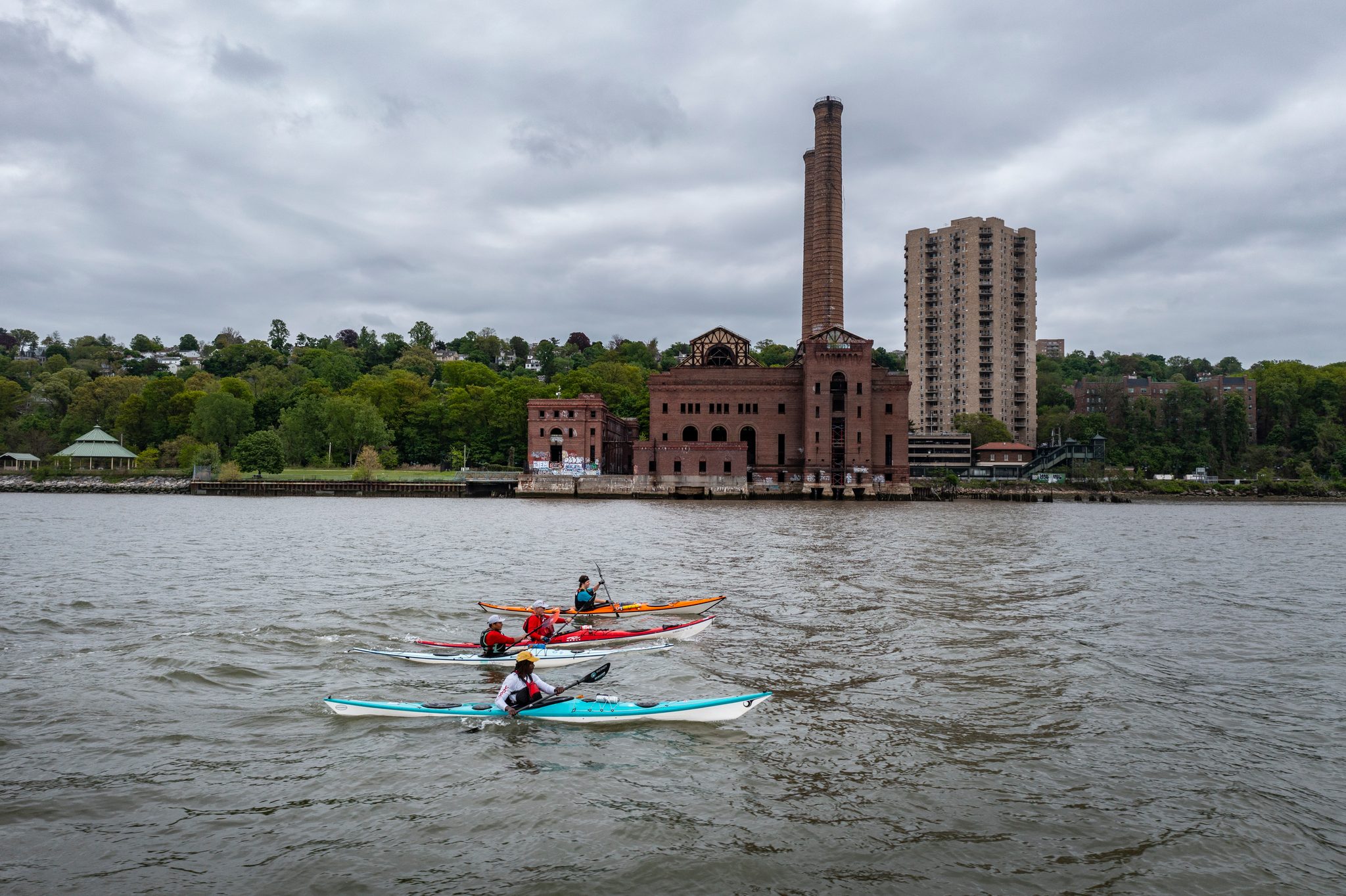 Kayaks slide past a brick power station and its tall smokestack, the city starting to crowd the shoreline.