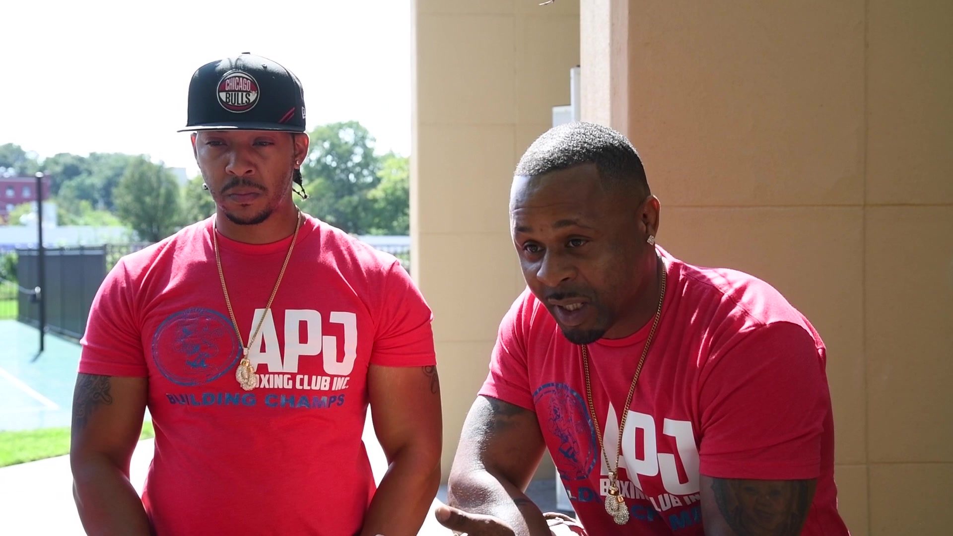 Outside the venue, Coach Kariym leans in and talks with his hands while a man in a Bulls cap listens beside him.
