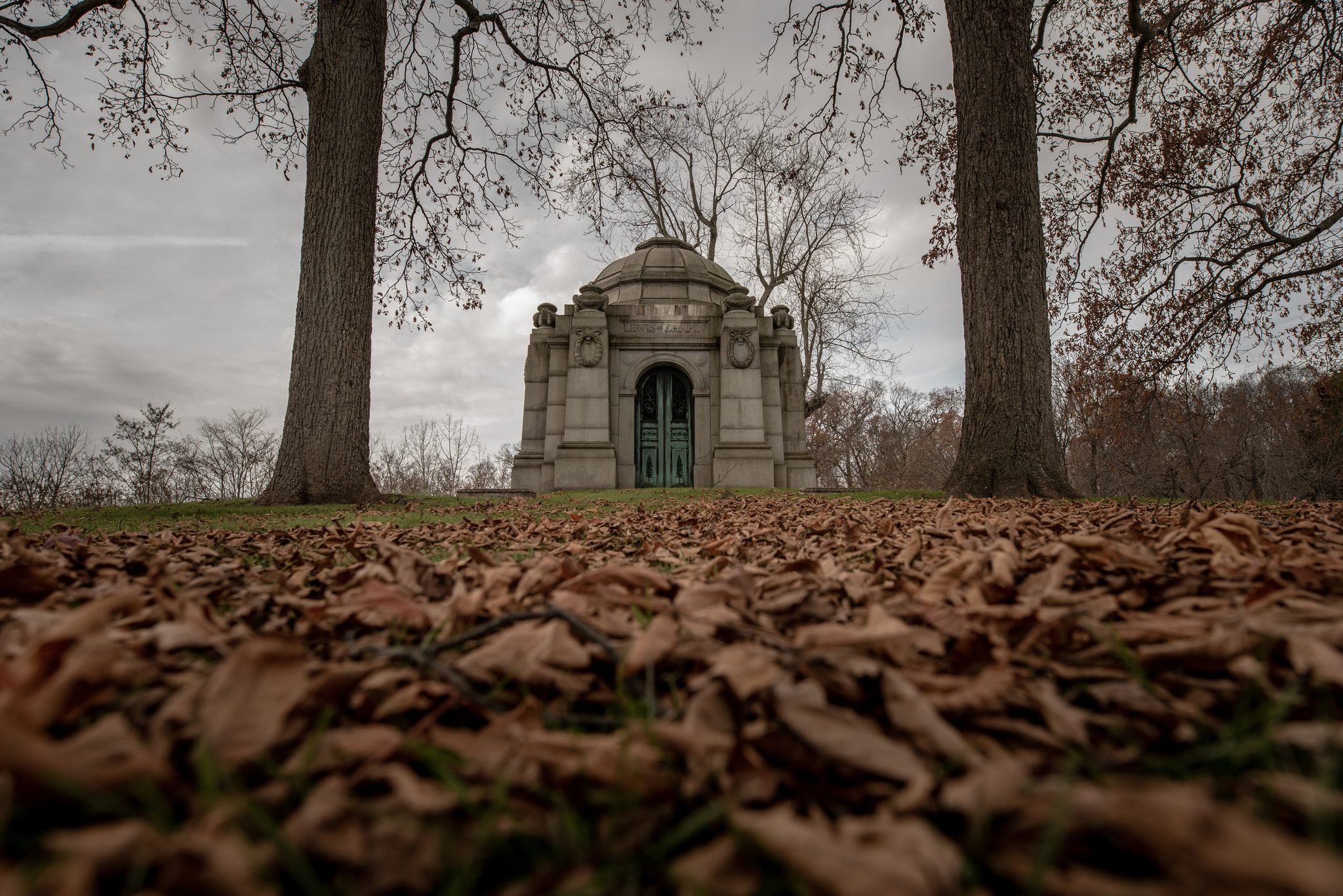 Fallen leaves carpet the ground between two old trees, the mausoleum waiting beyond.