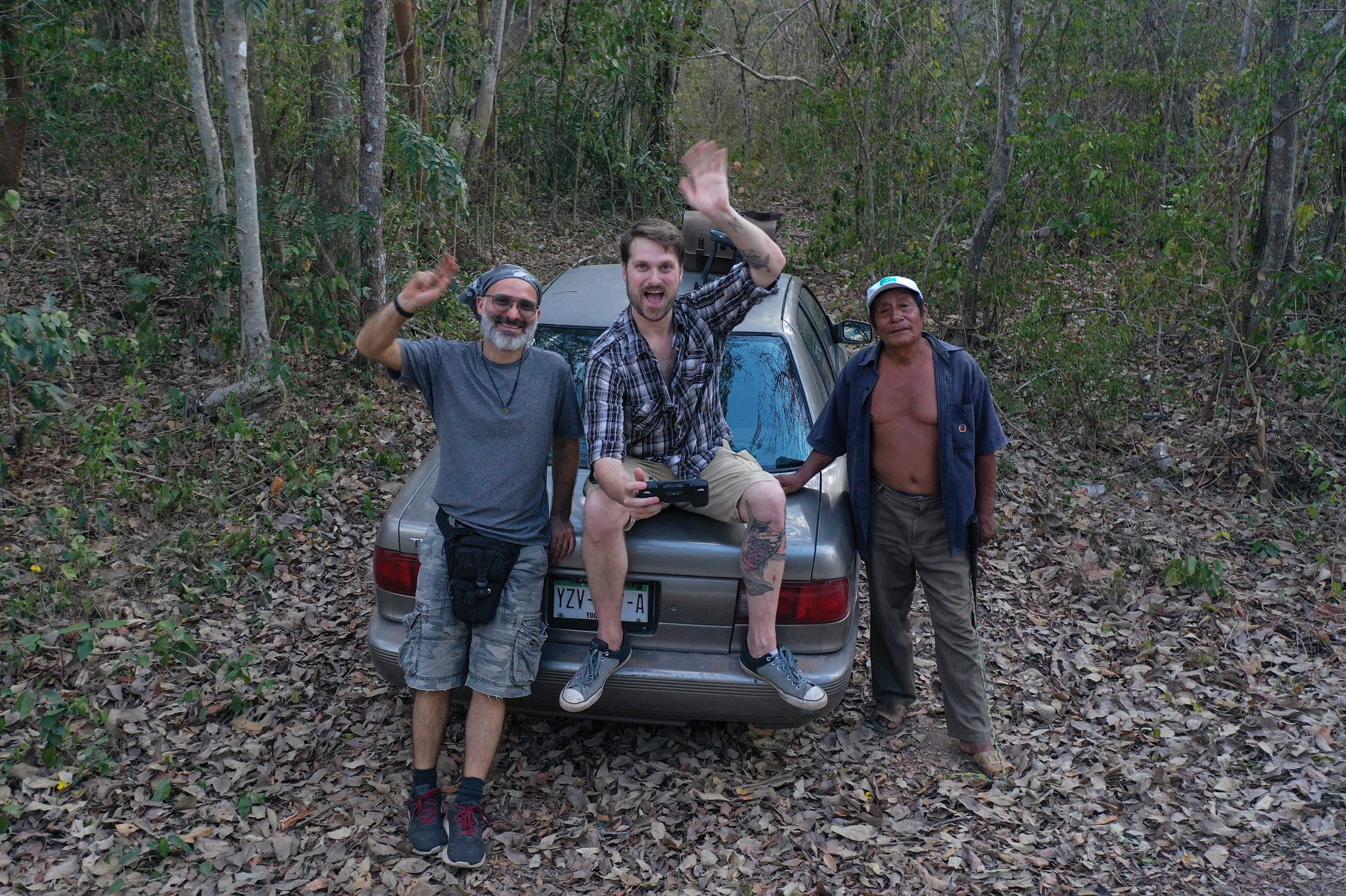 Three men wave from the trunk of a car in the forest.