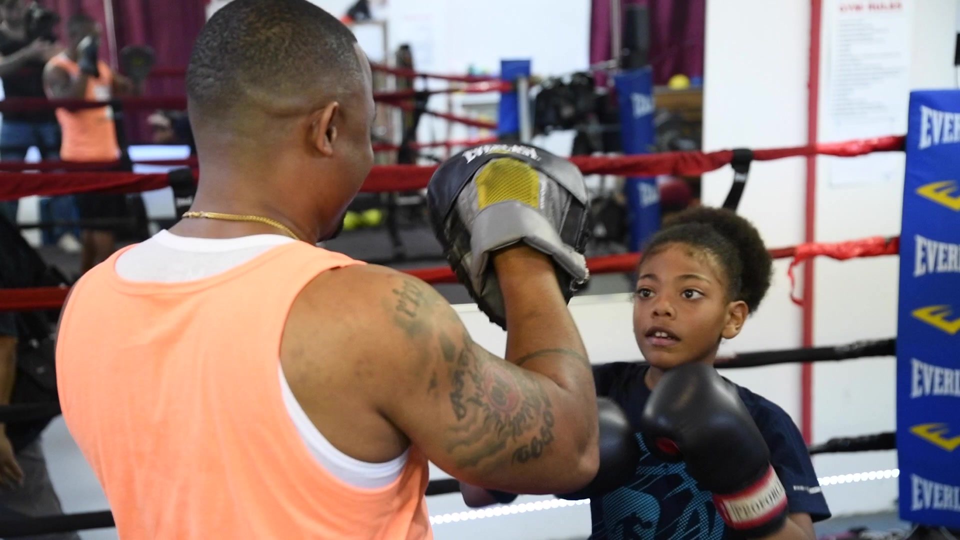 A young boxer watches the pads closely, gloves up, while the coach sets the next target.