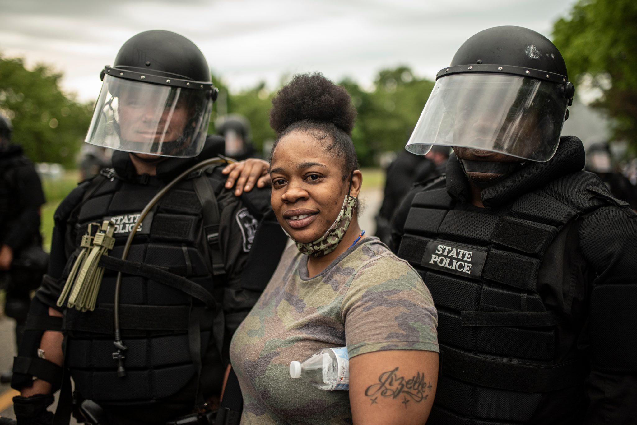 A woman smiles between two troopers in riot gear, one of them resting a hand on her shoulder.