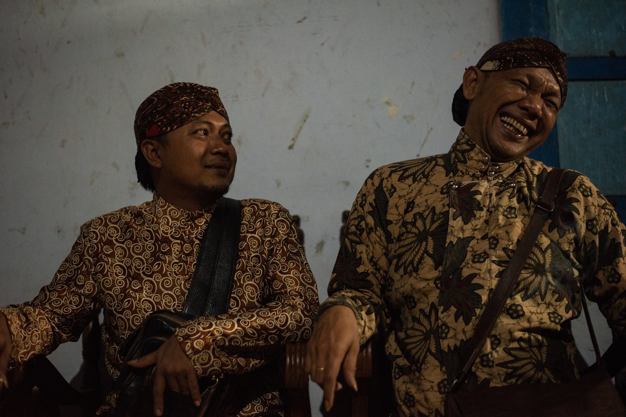 Two men in batik and headcloths share a laugh against the plaster wall.
