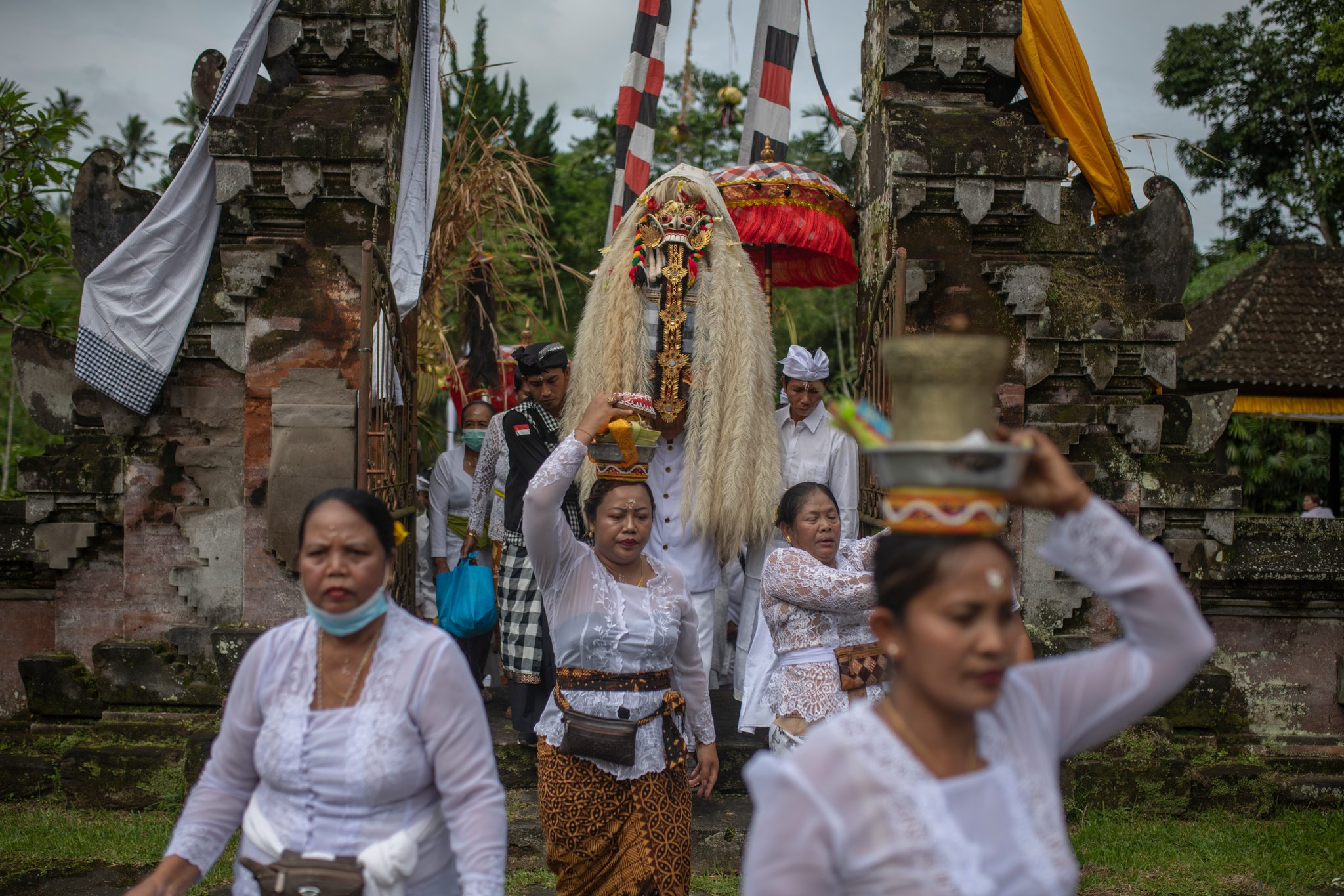 Women carry offerings through the split stone gate, the masked figure close on their heels.