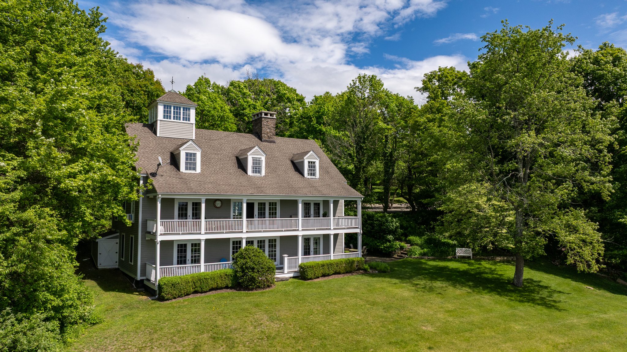 Double porches and a rooftop cupola on a gray clapboard colonial, set among lawns and late May trees.