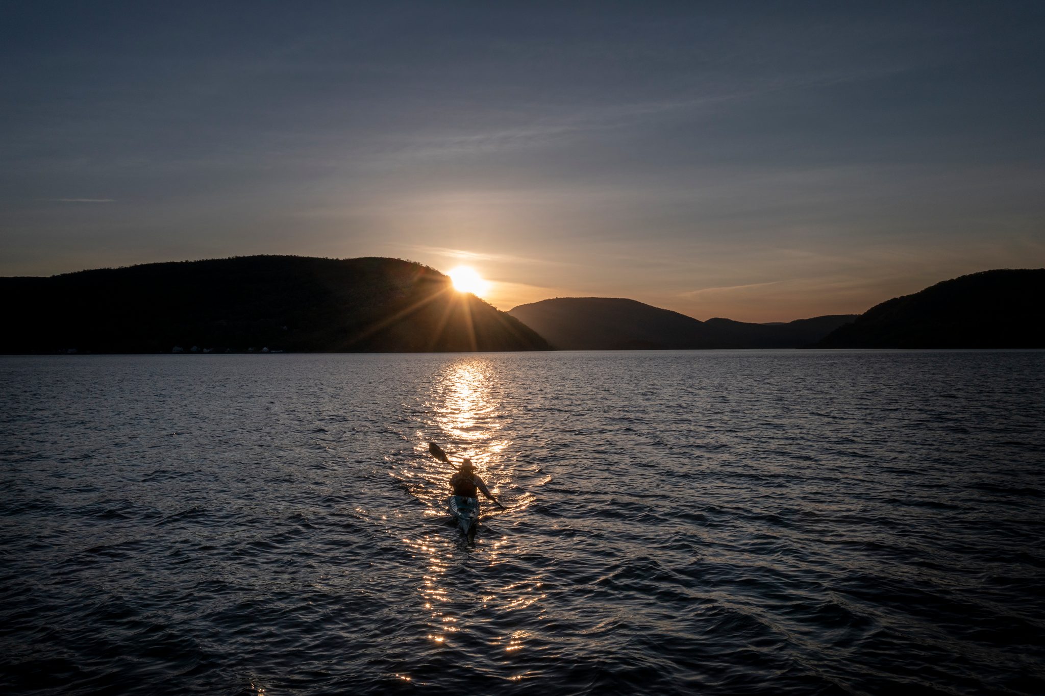 The sun drops behind the ridge and a single paddler rides the gold streak it leaves across the river.