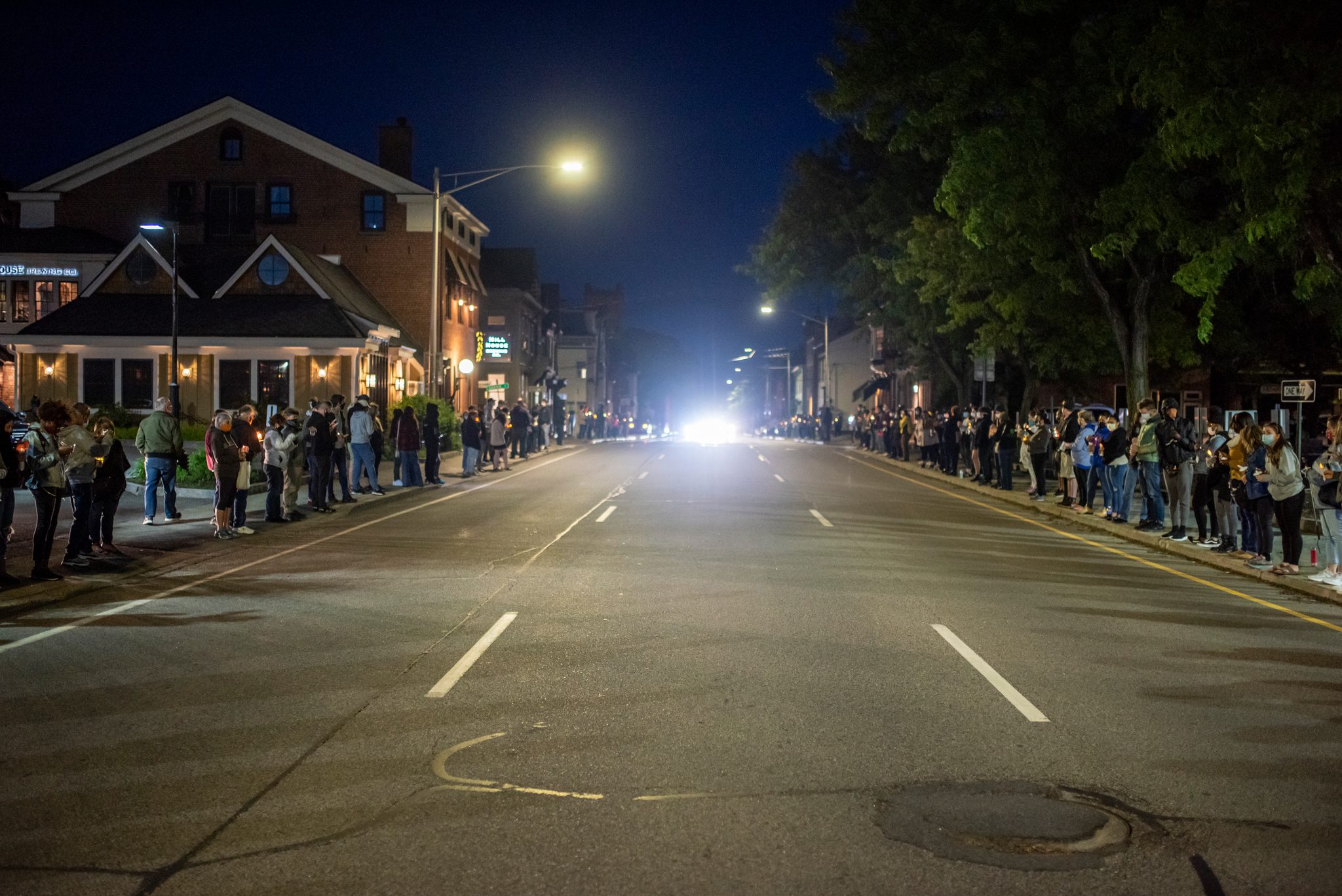 People line both sides of an empty street by candlelight, a single bright light waiting at the far end.