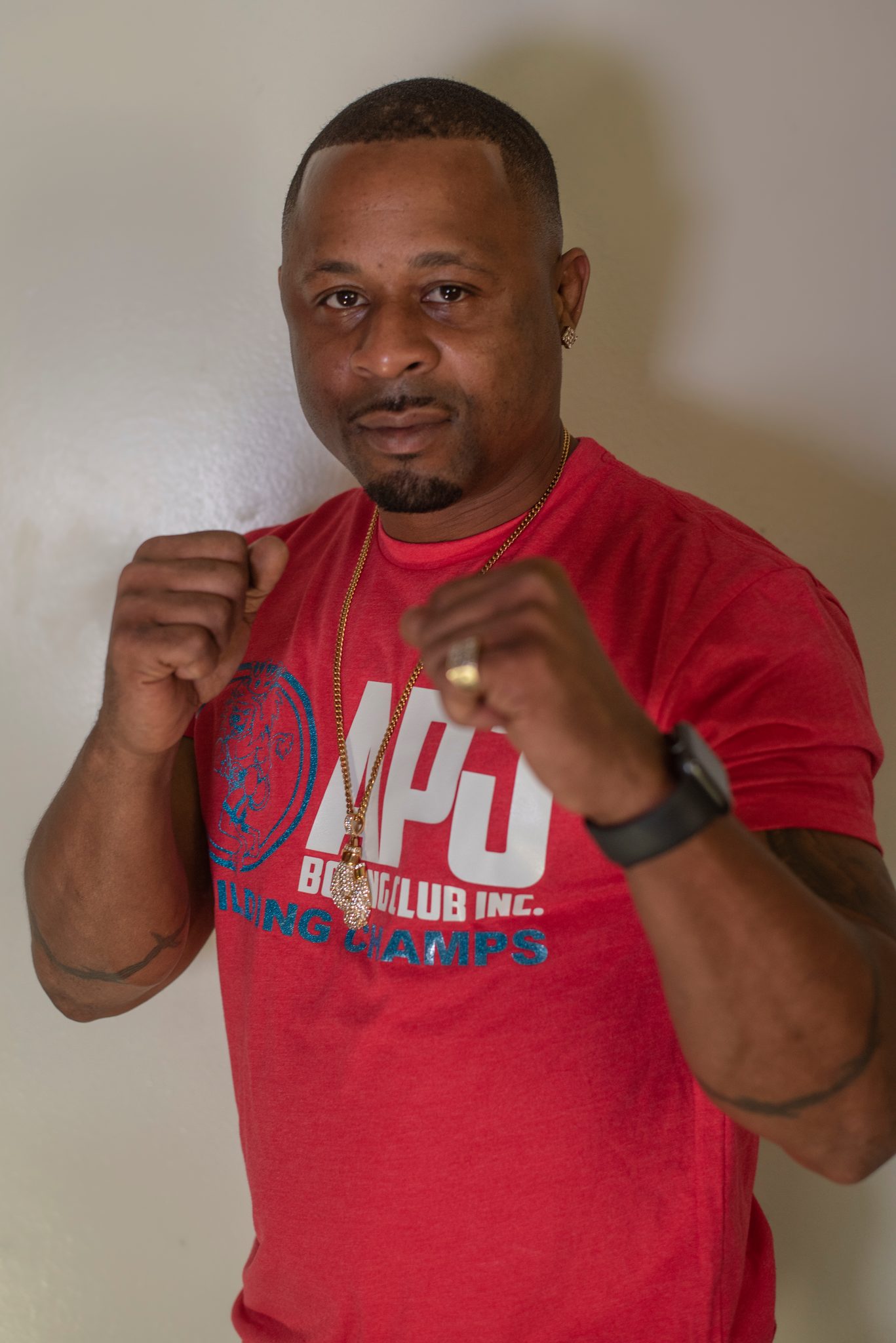 Coach Kariym brings his fists up against a plain wall, a watch on one wrist and a gold chain over the club shirt.