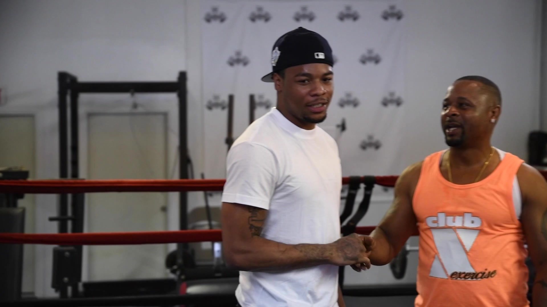 A handshake by the ring between a man in a white tee and Coach Kariym in an orange Club X tank.
