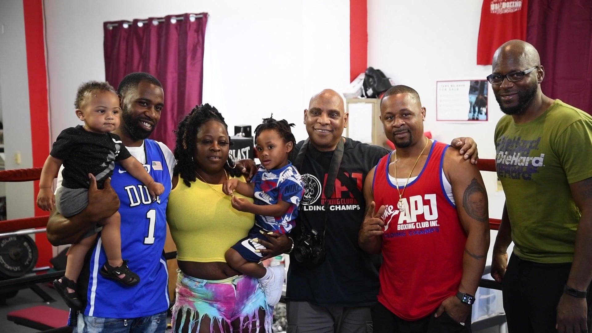 A group gathers for a photo by the ring, two toddlers held up in arms and Coach Kariym in a red club tank.