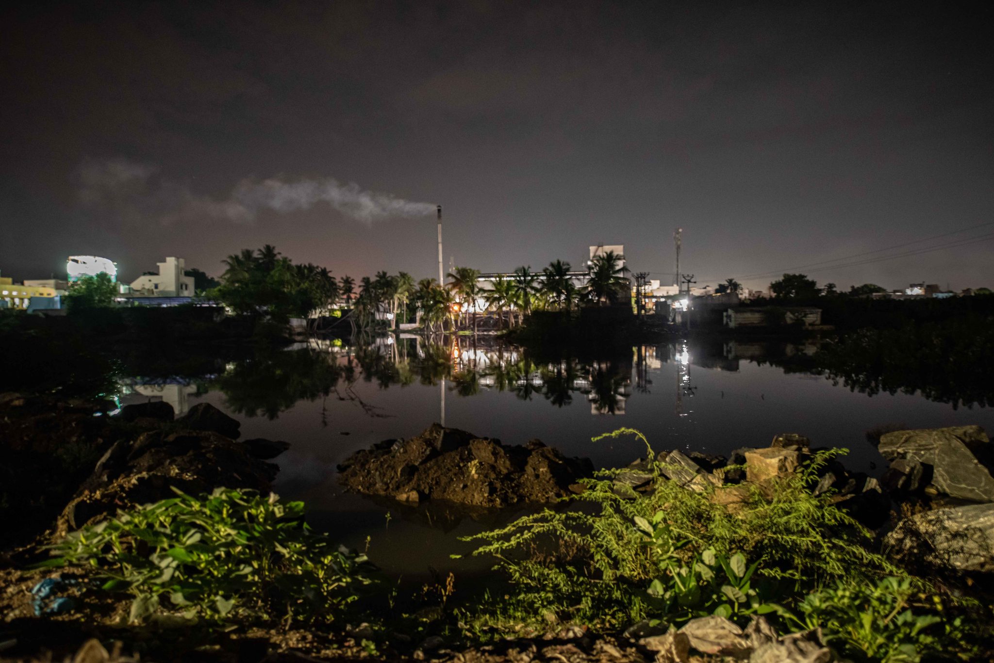The lights of the town and a smoking chimney reflect in the still river at night.