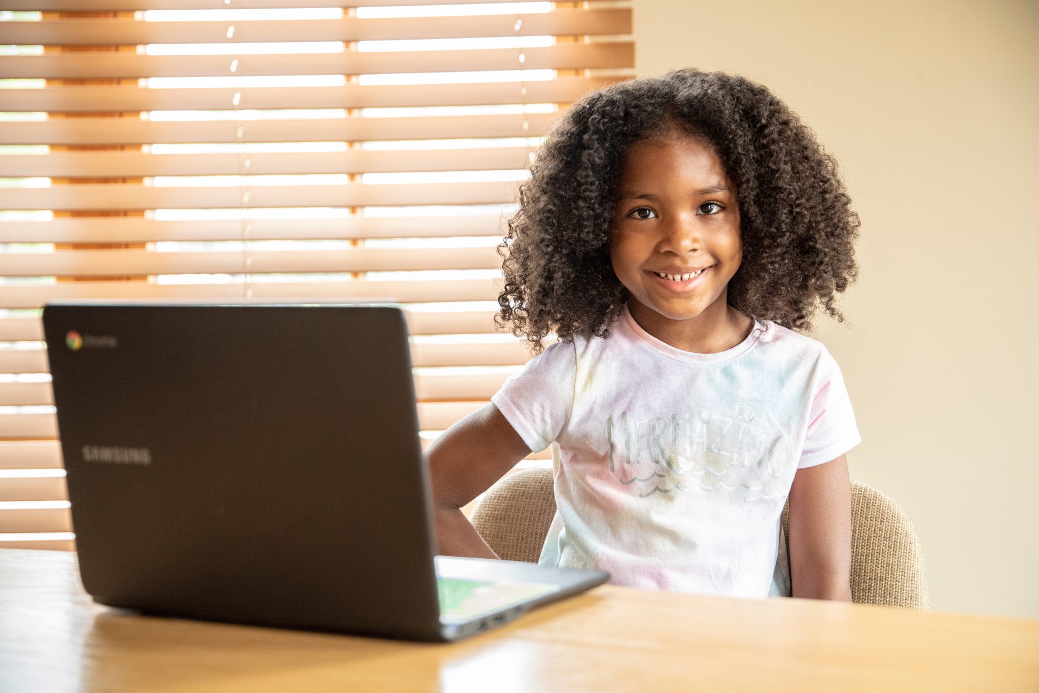 Laptop open, a young girl smiles for the camera at the kitchen table.