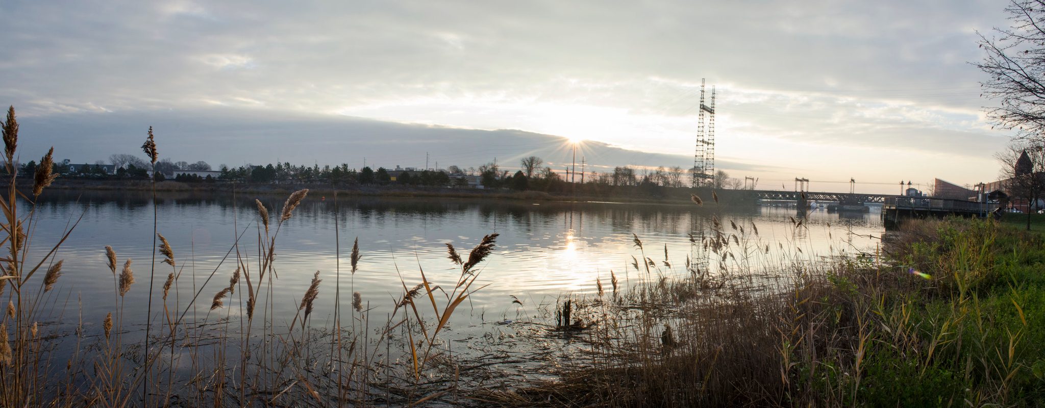 Across the river marsh, a bridge and a tower stand against the setting sun.