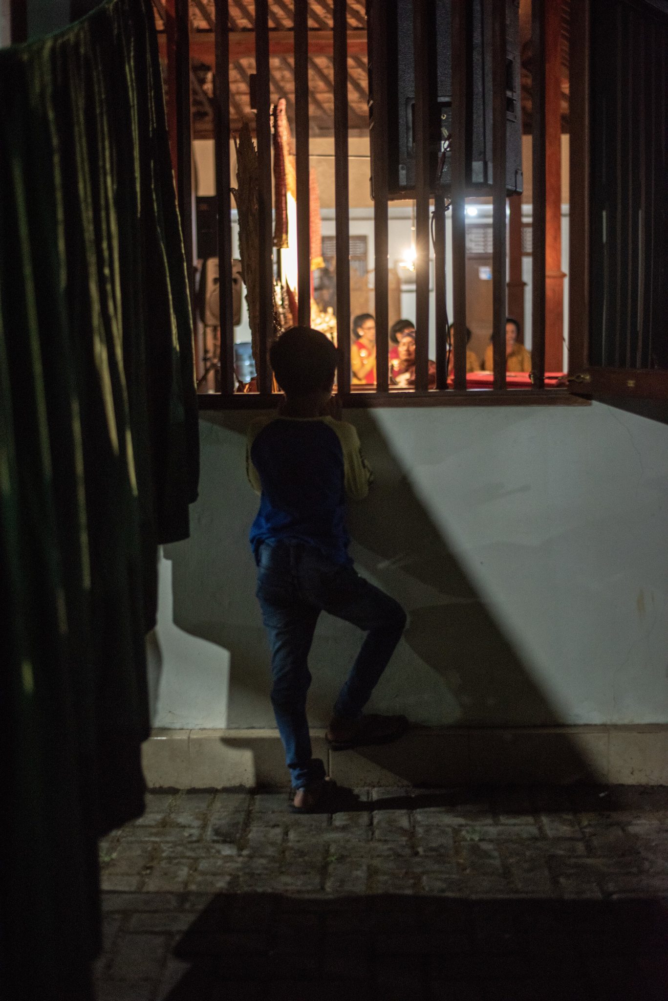 A boy props a foot on the wall outside to see through the window bars.