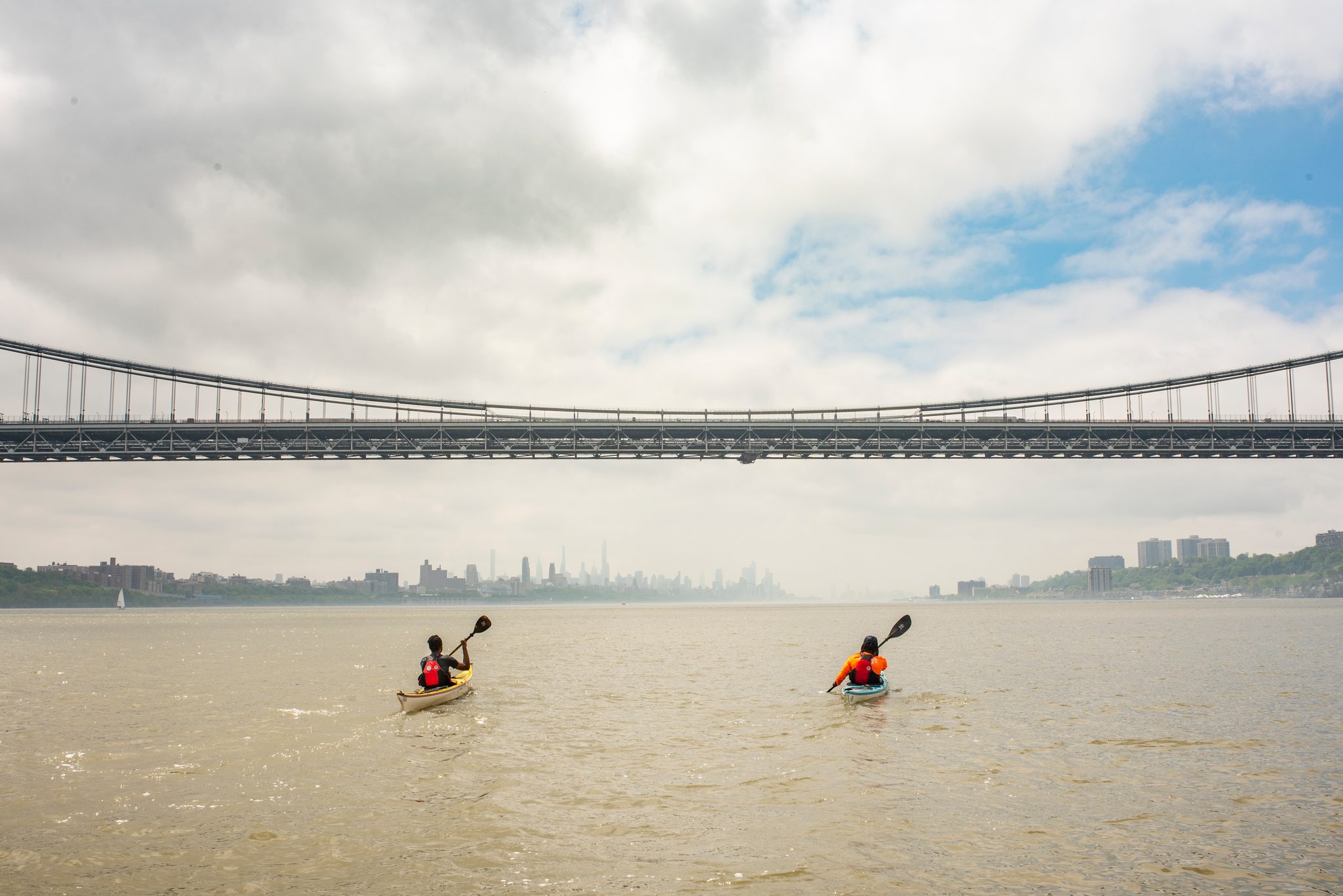 Two small figures paddle toward the long span of the bridge, the skyline a pale smudge beyond it.