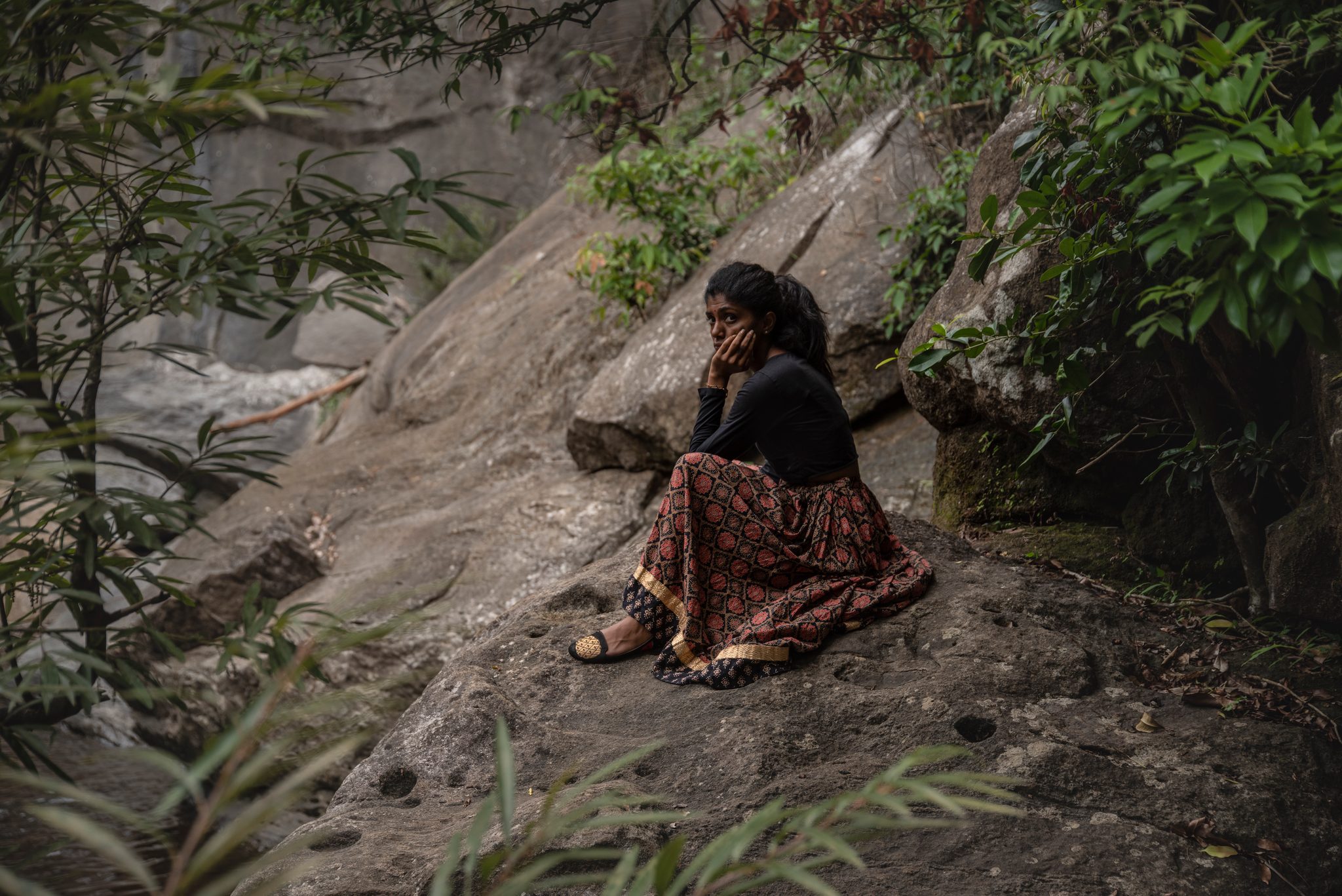 A young woman sits on the rock face, chin in hand, the forest all around her.