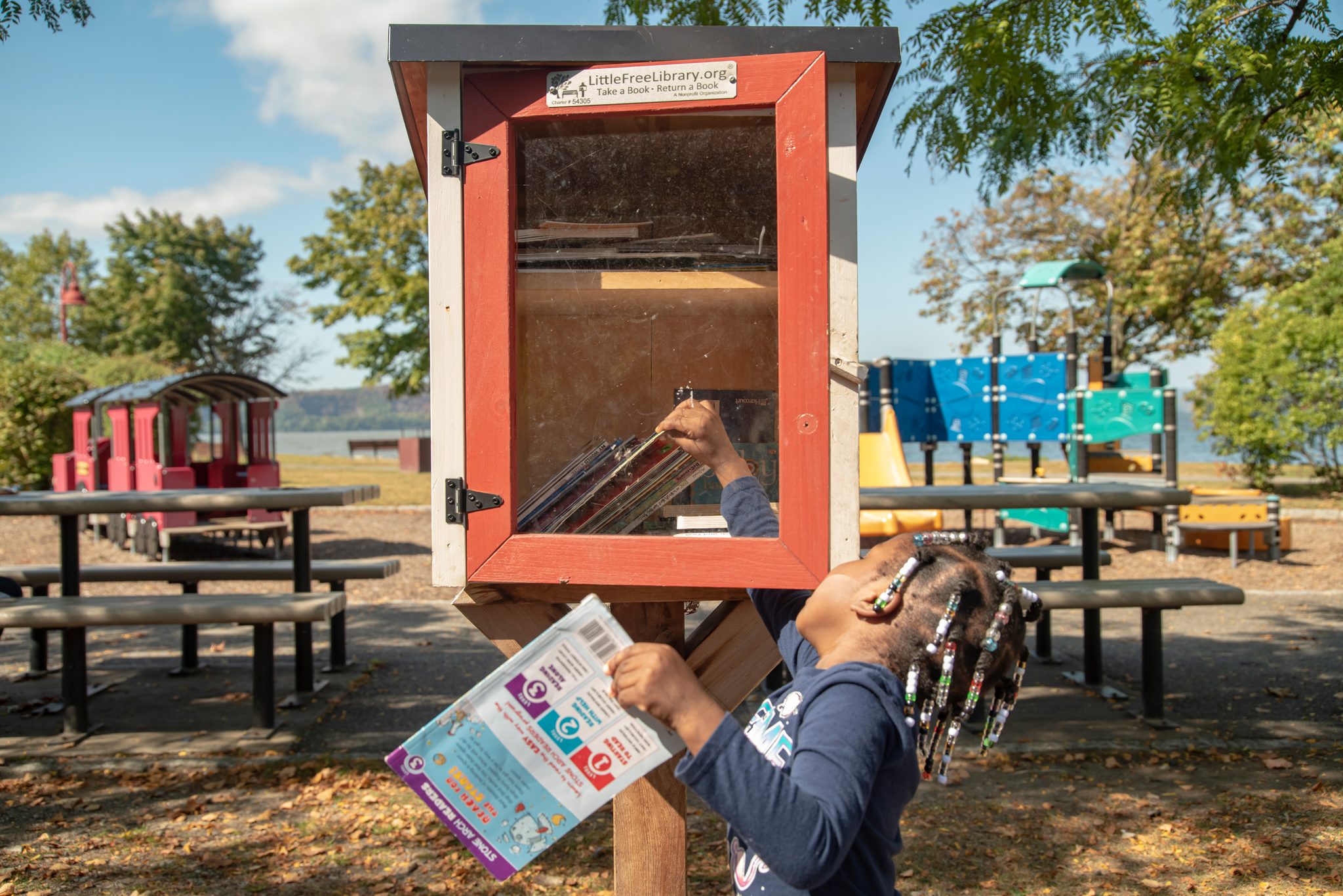 A girl with beaded braids reads right there at the Little Free Library, too curious to carry the book home first.