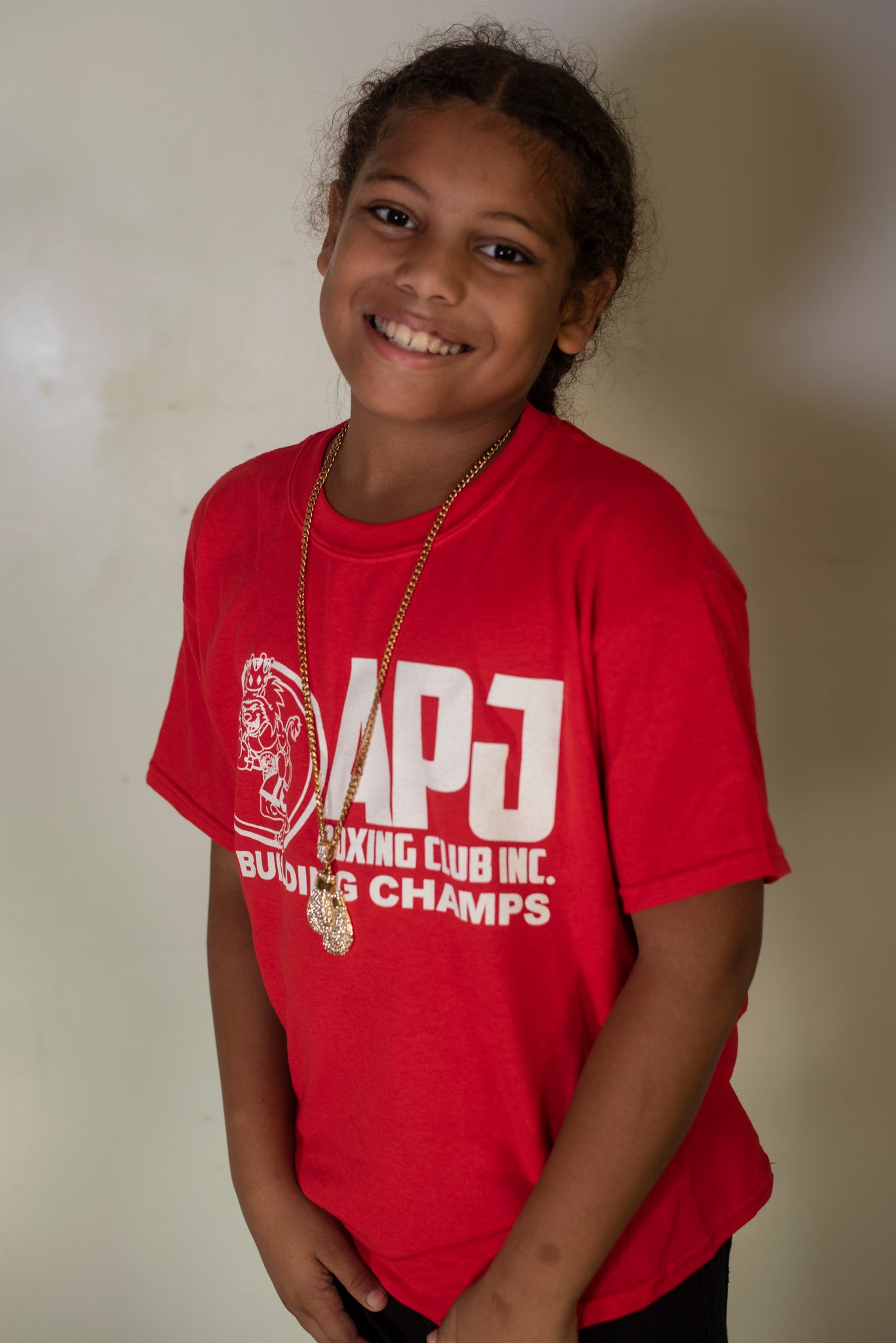 A girl with braided hair and a gold chain grins wide, head tipped toward her shoulder.