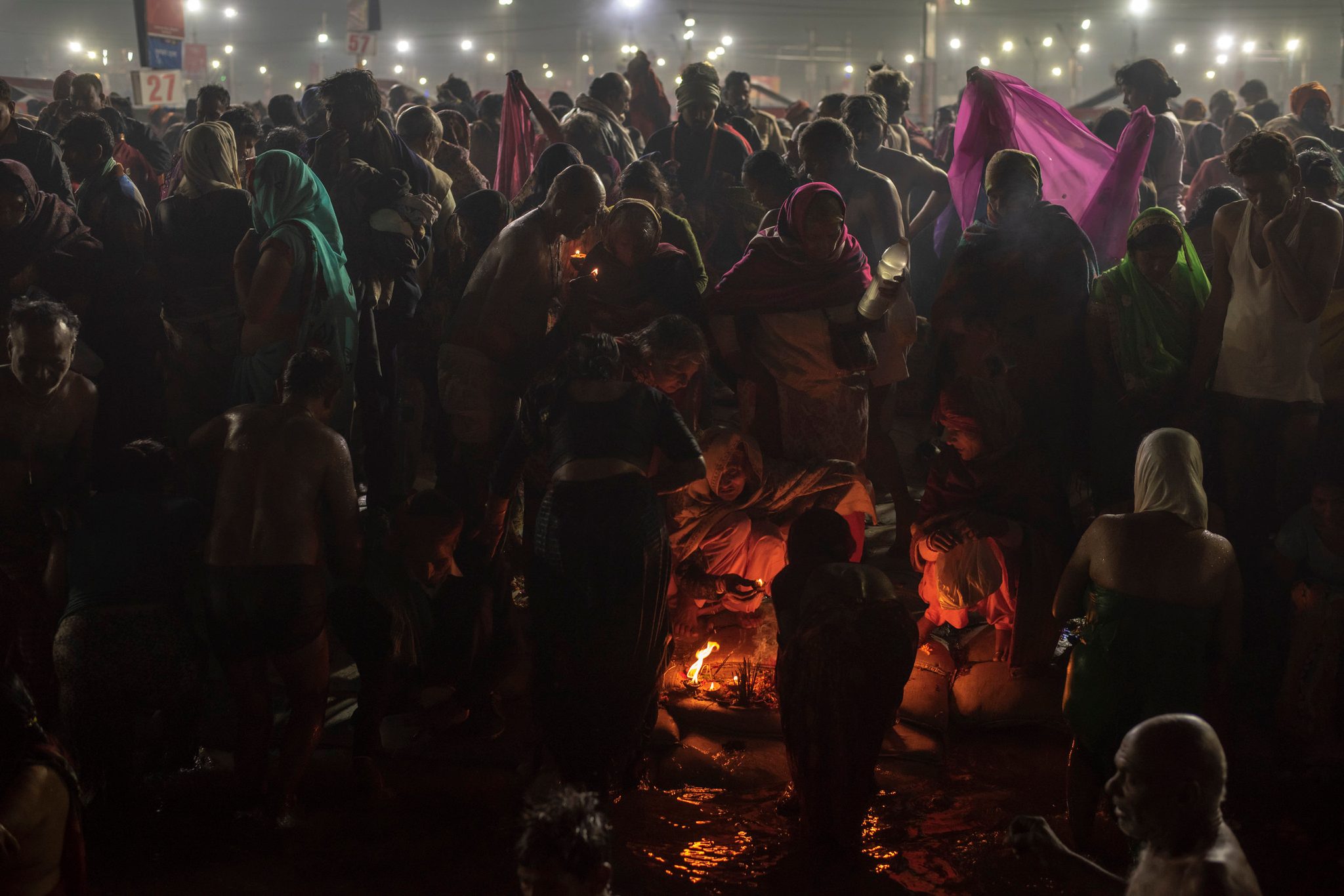 Thousands press toward the water through the night, a fire glowing at the heart of the crowd.