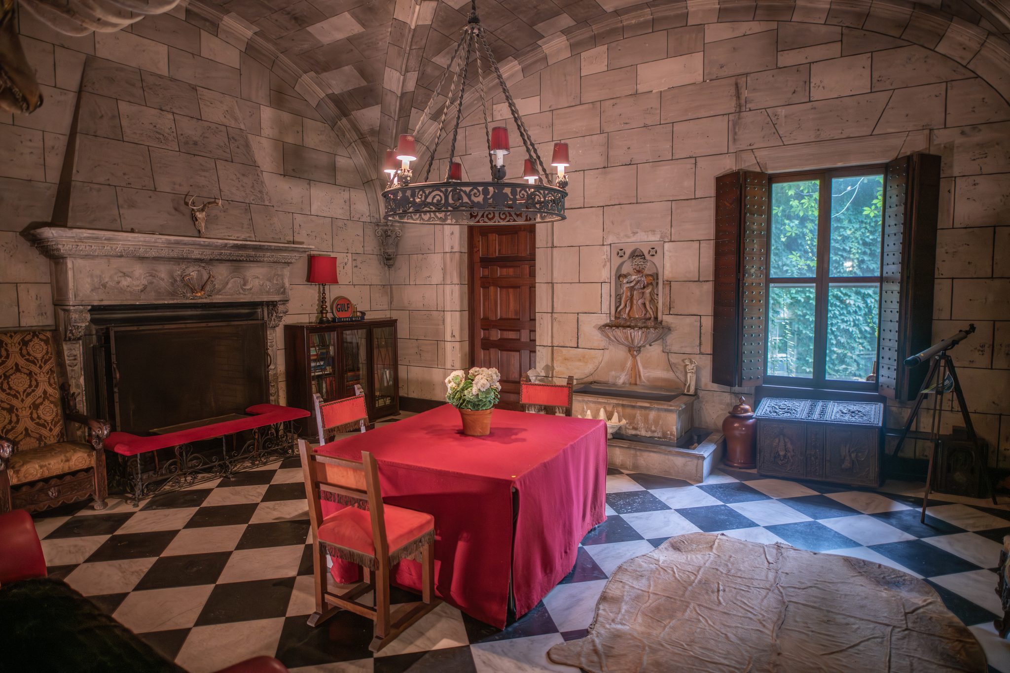A stone-vaulted room with a carved stone fireplace, a checkerboard floor, an iron chandelier and a brass telescope by the window.