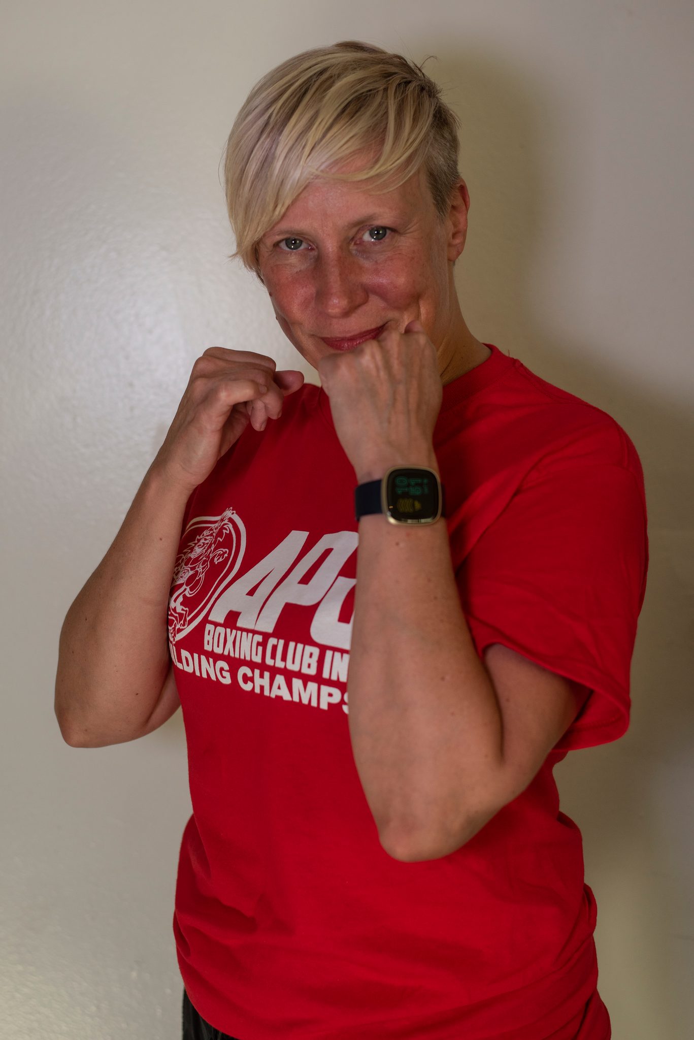 A woman with short blonde hair grins behind her fists, a smartwatch on her raised wrist.