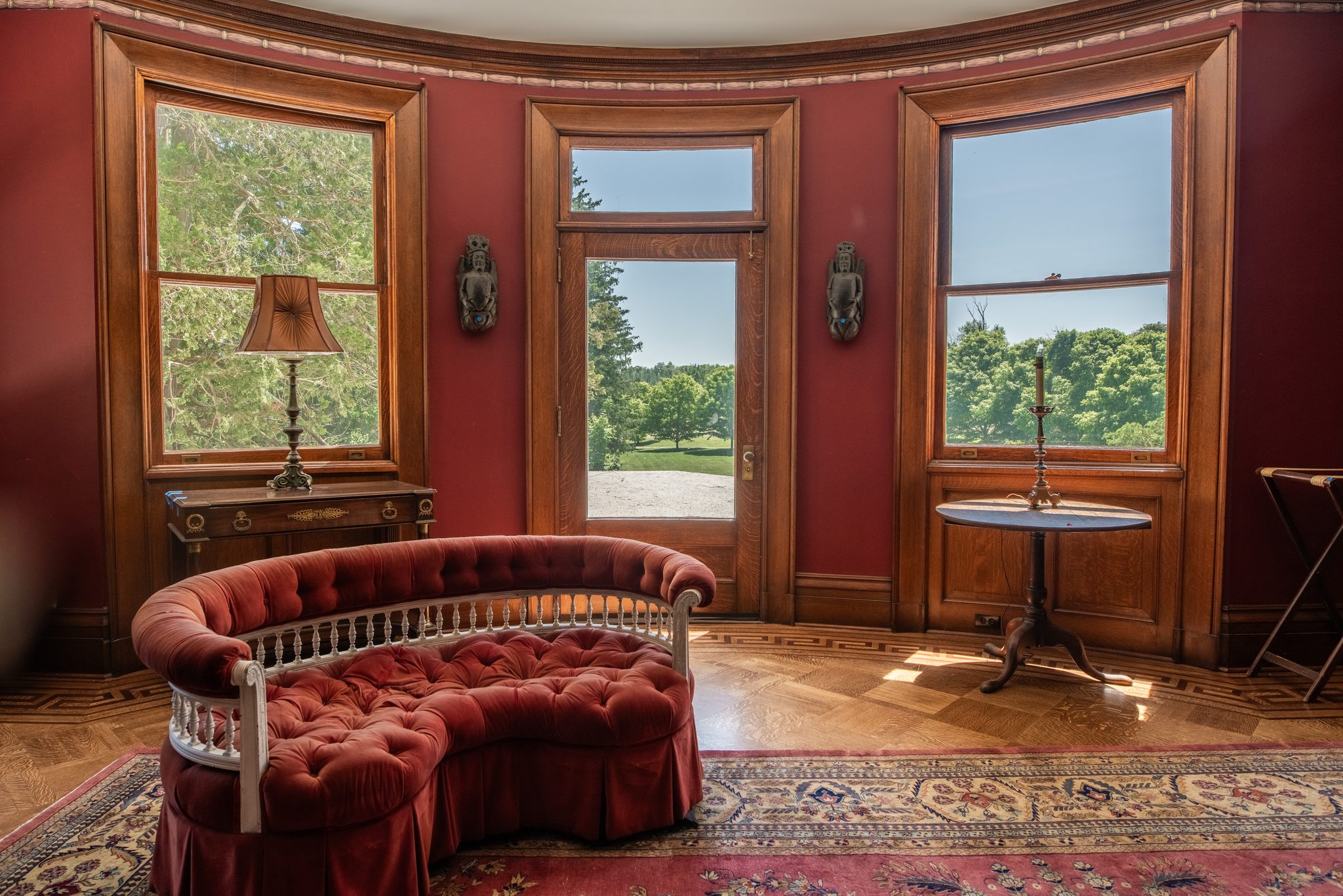 A red-walled round room with oak window casings, a tufted velvet settee and a door opening onto the porch roof.