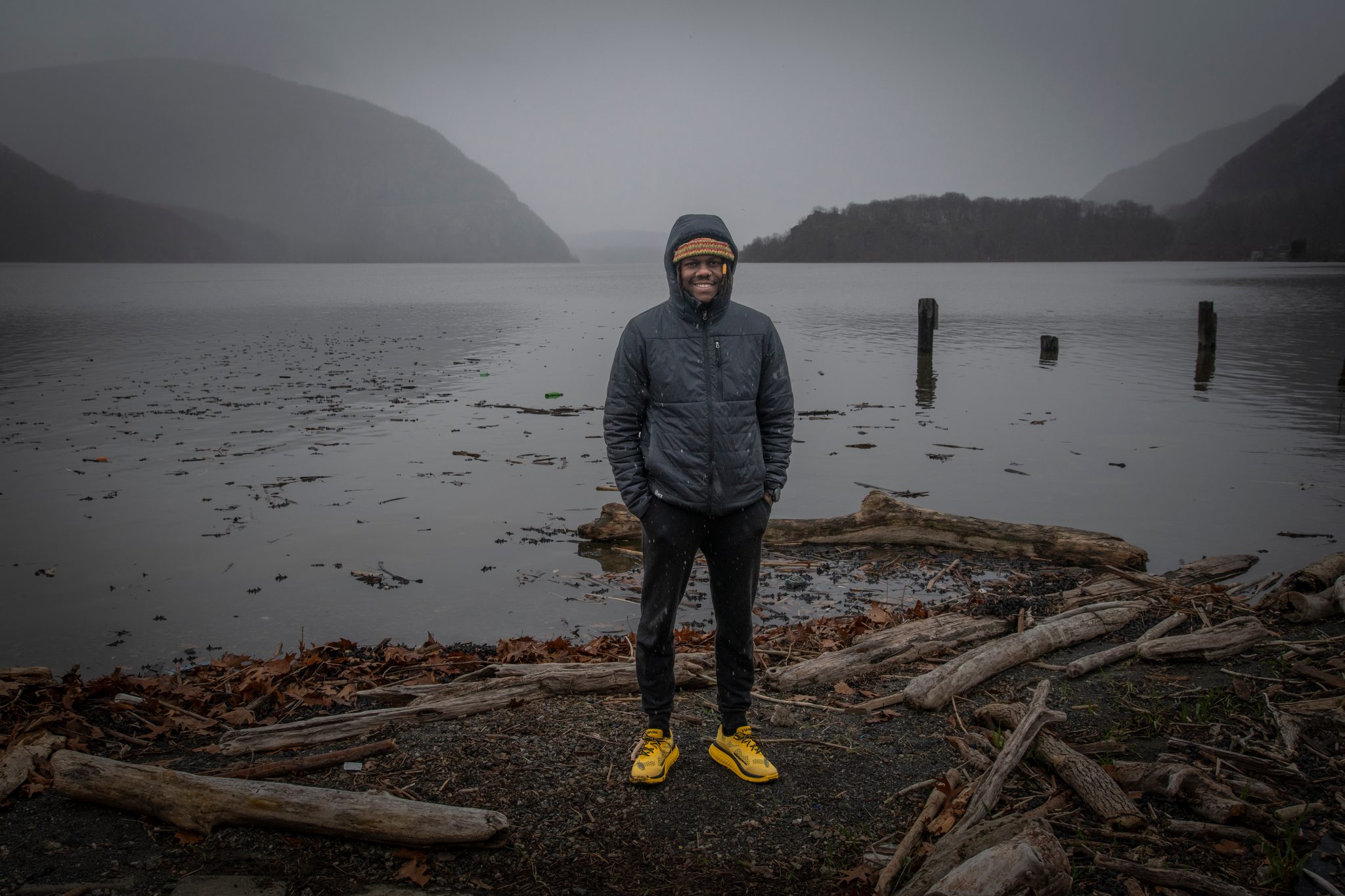 Chev stands on a driftwood shore in grey drizzle, yellow shoes the only colour against the river and mist.