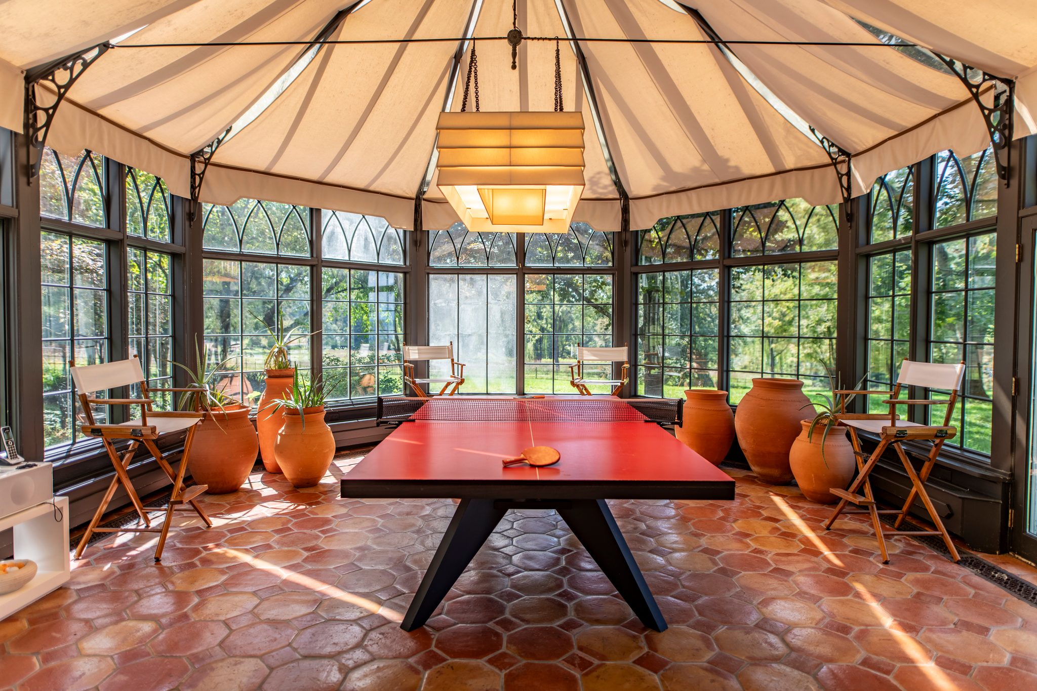 A tented glass conservatory glows at midday, terracotta hex tiles and clay urns ringing a bright red table.