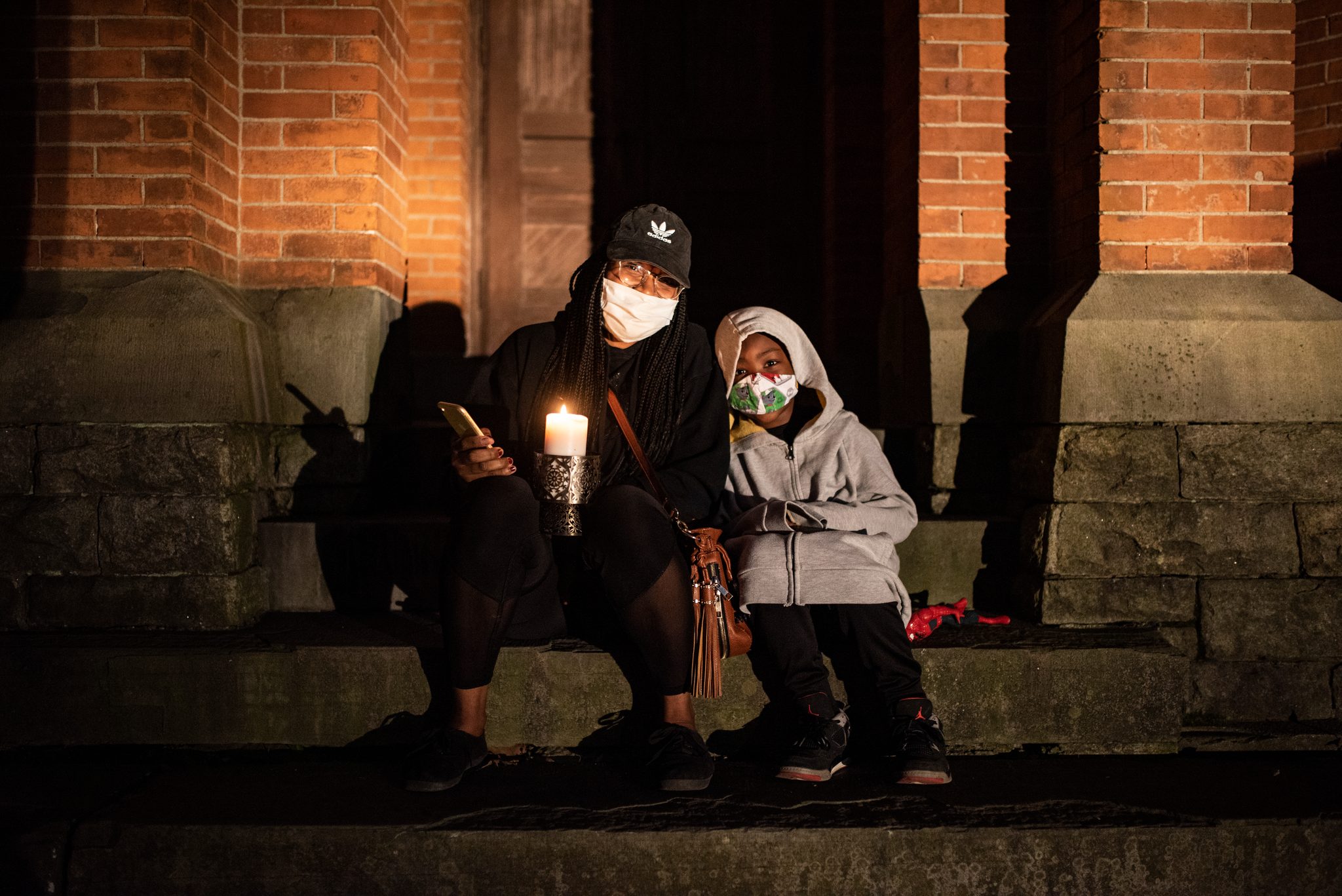 Two mourners sit on the church steps, candles in hand, masks on.