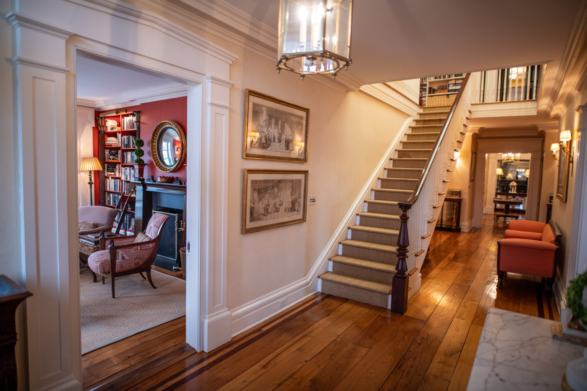 Wide plank floors with an inlaid border run past a long staircase, a red library glowing through the doorway in December.