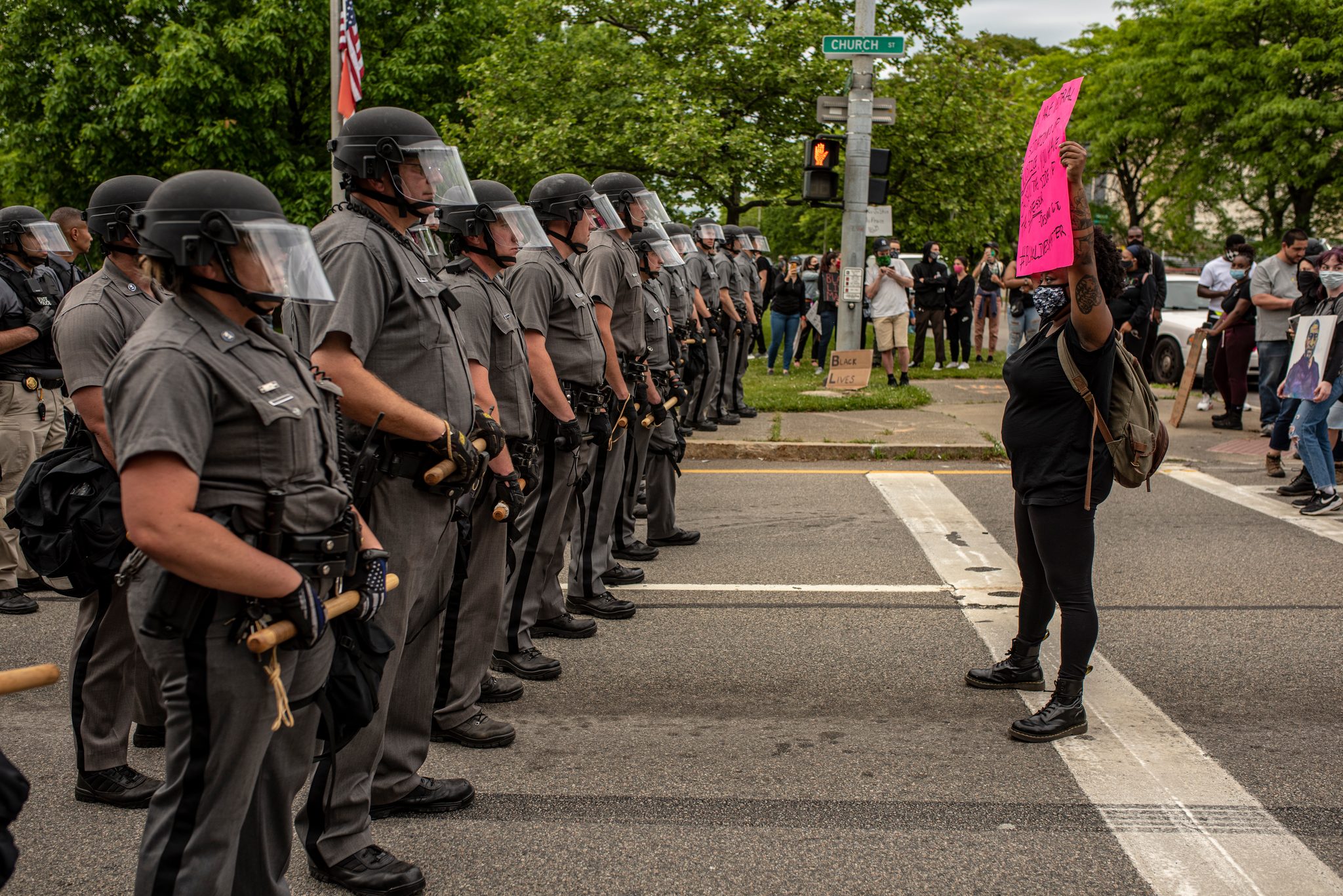 A woman stands alone in the crosswalk facing a line of troopers, her pink sign held high.