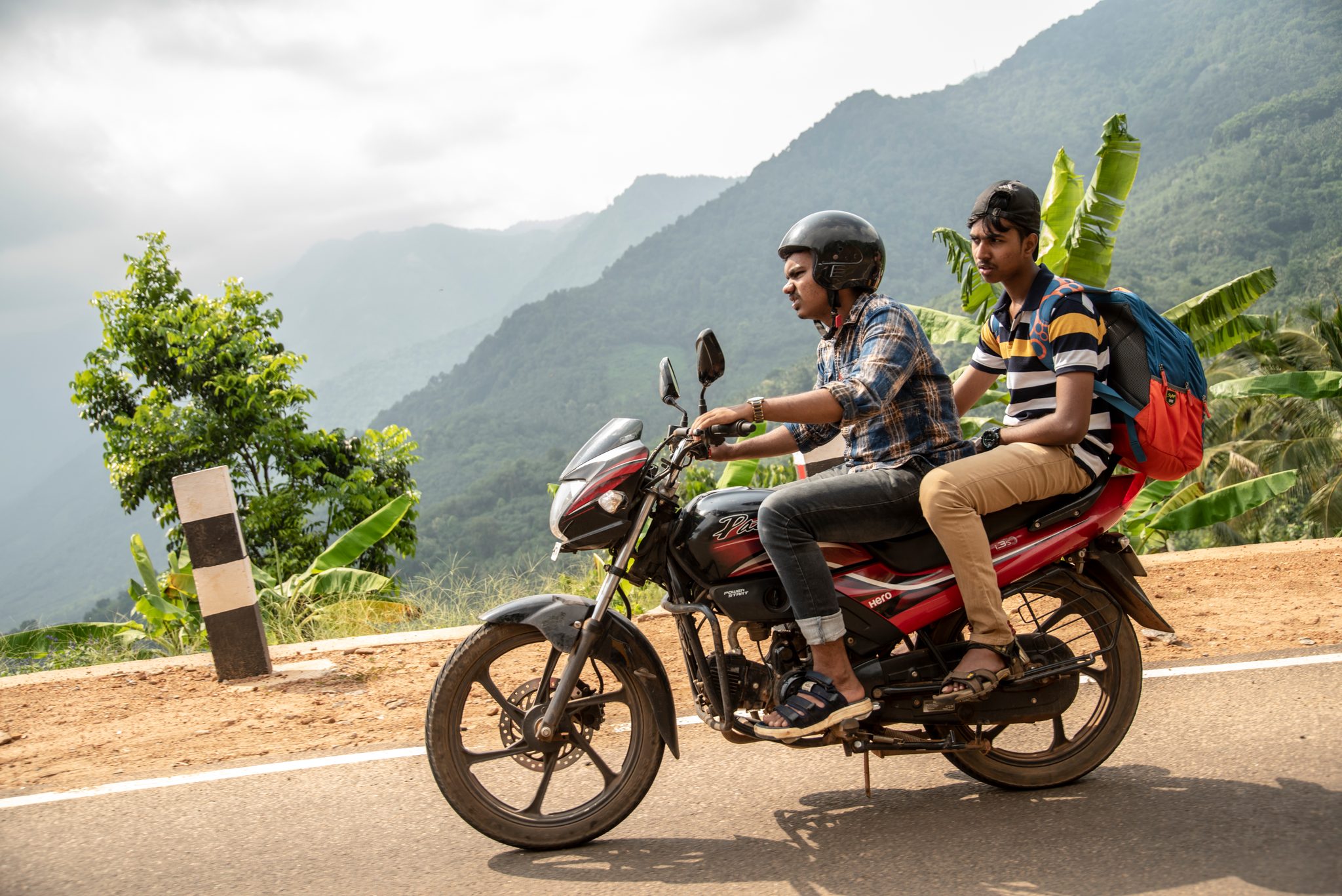 Two young men ride a motorcycle along the mountain road, the valley falling away behind them.