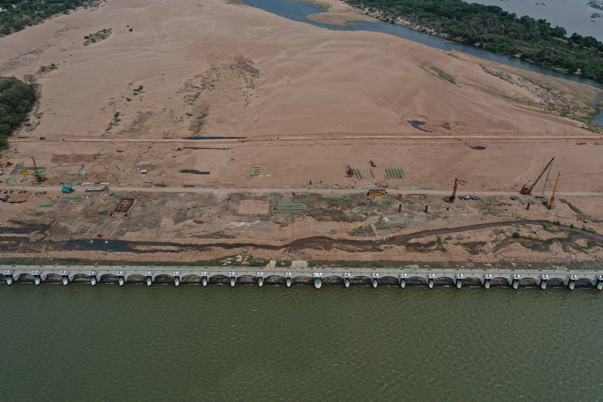 Construction crews work the sand beside a long bridge over the river.