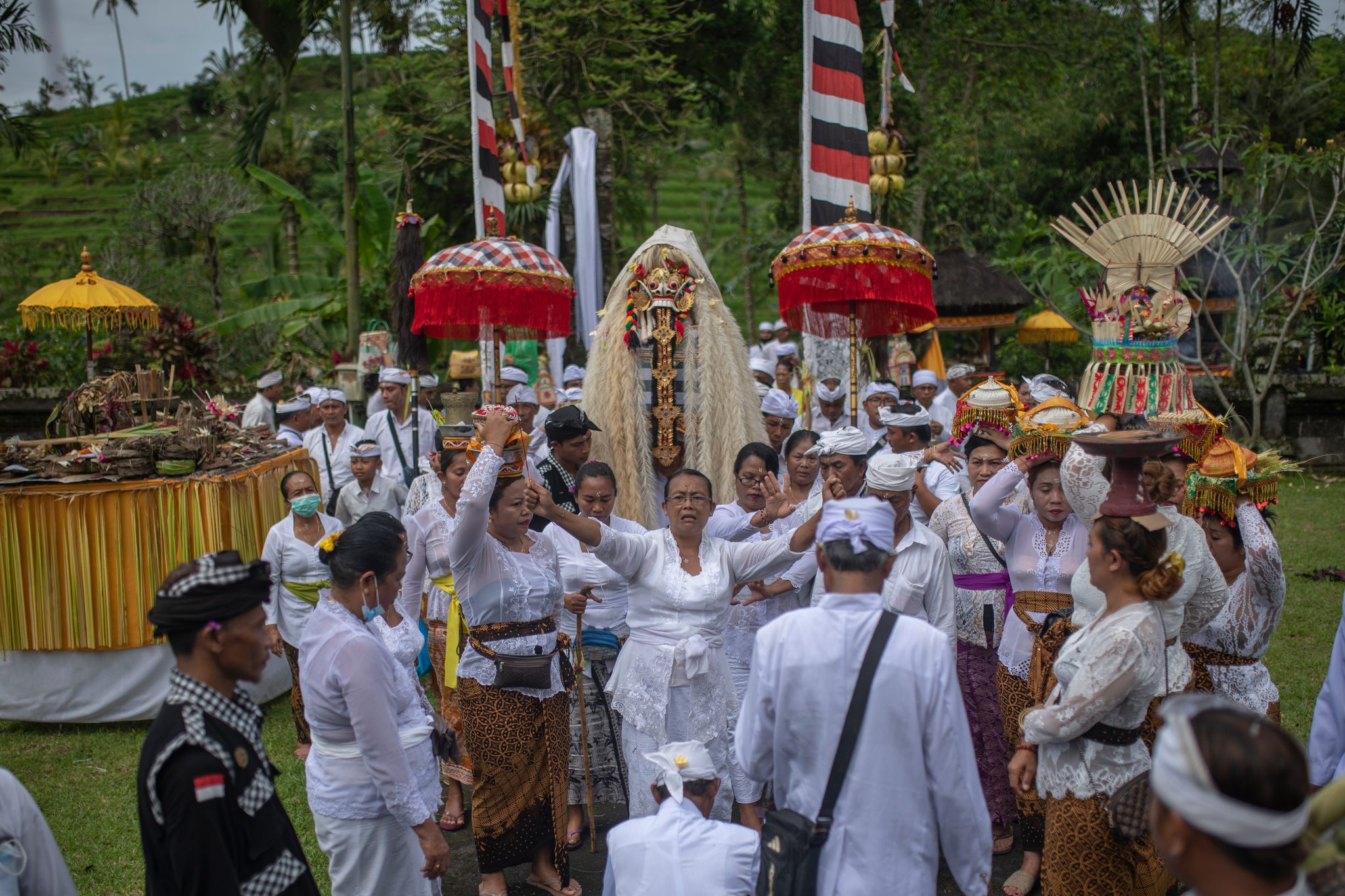 The procession moves out beneath fringed parasols, offerings balanced on heads and a masked figure towering over the crowd.