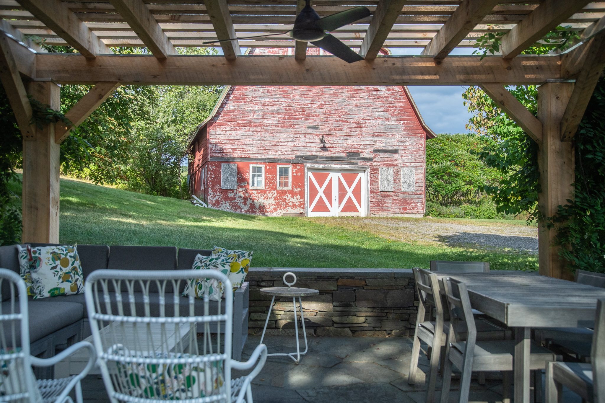 Under a timber pergola in late August, an old red barn with white cross-braced doors fills the view across the lawn.