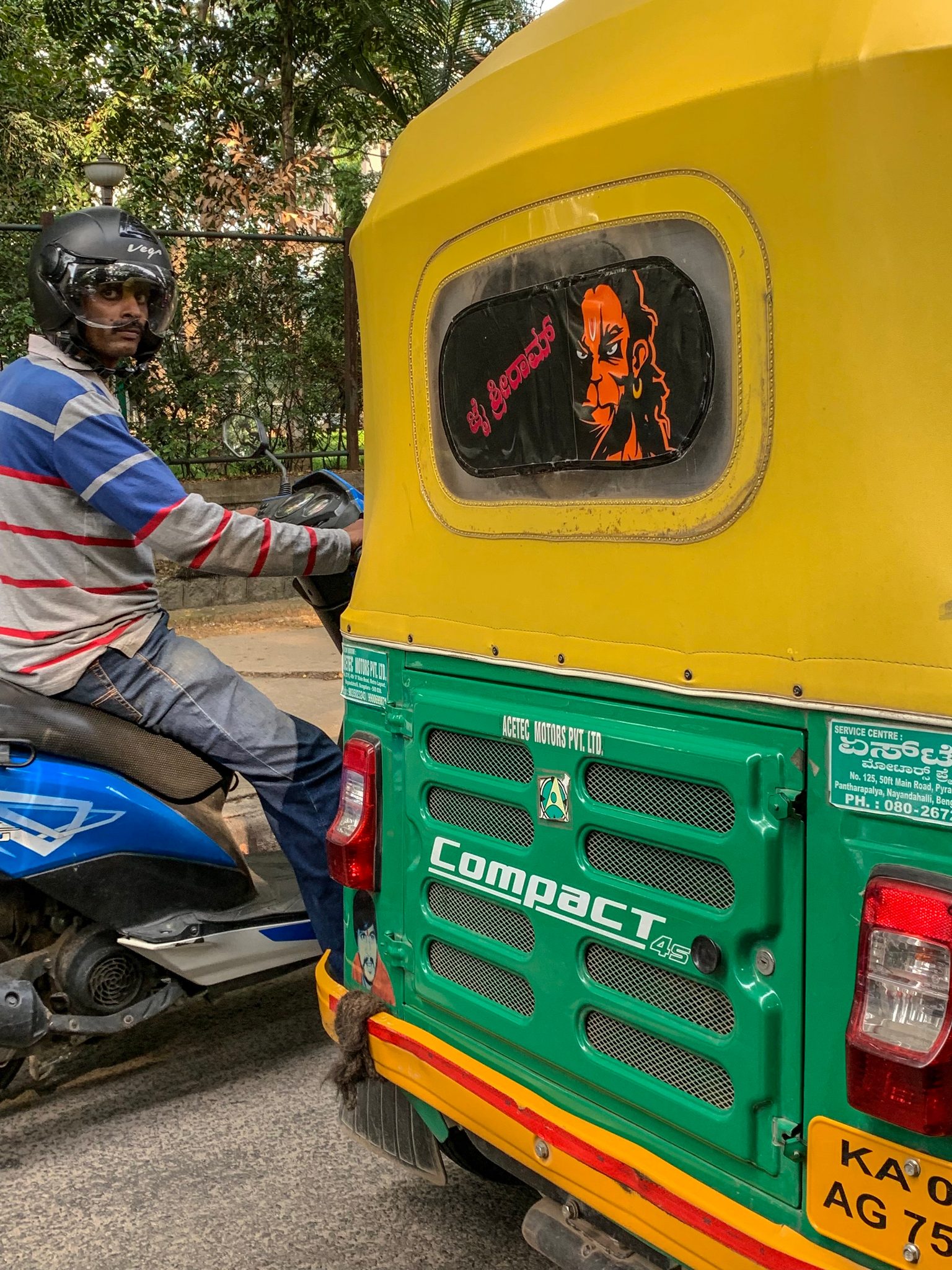 A rider on a scooter waits beside a yellow and green auto rickshaw in traffic.