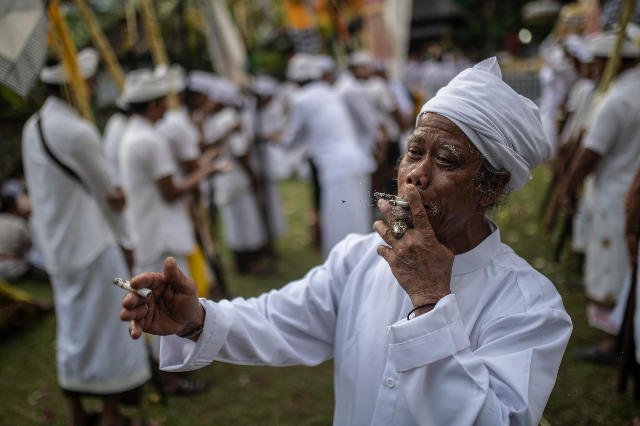 An elder in white, cigarette in hand, lets the ceremony swirl around him.