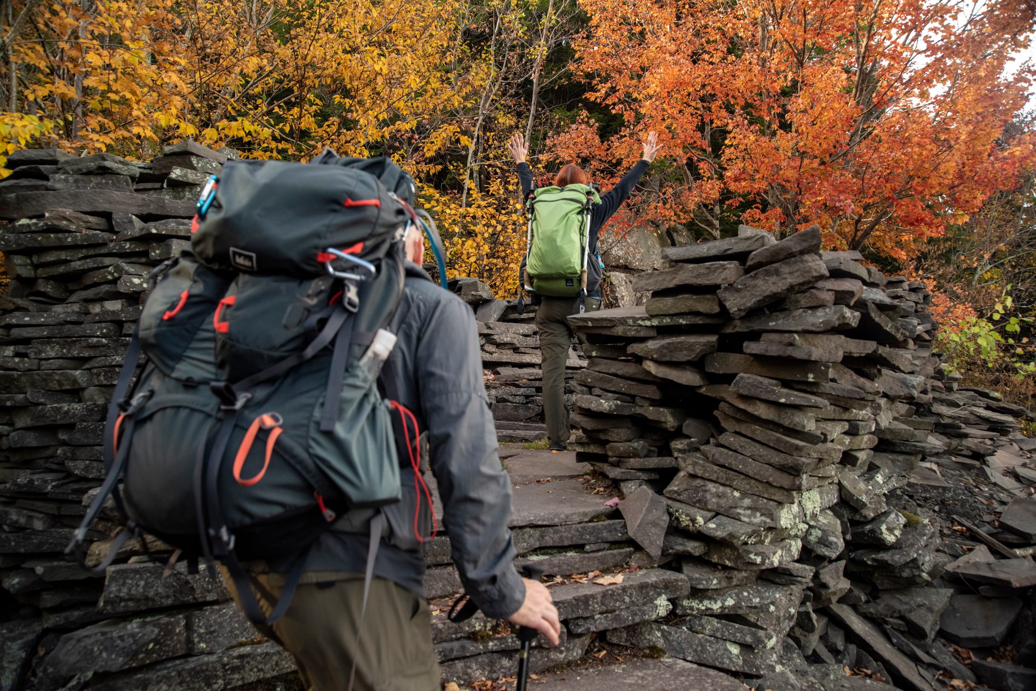 Backpacks climb through stacked slate and blazing autumn leaves, and the one out front throws his arms up at the top.
