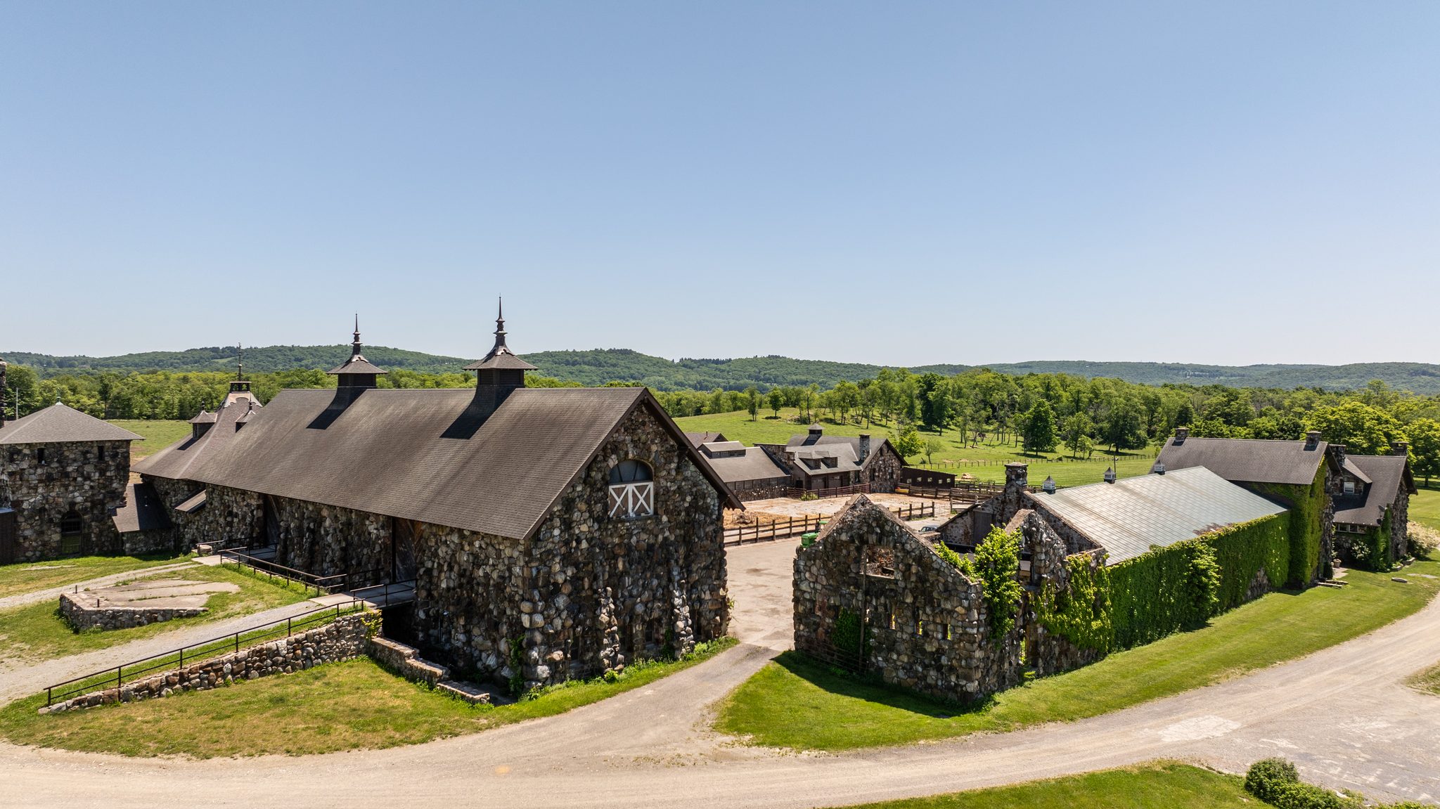 Fieldstone barns with steep roofs and spired cupolas, a gravel lane curving past them toward open pasture.
