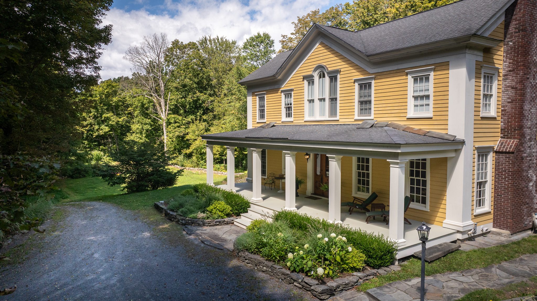 A butter-yellow Greek Revival farmhouse with a deep columned porch, its gravel drive curving in under late summer trees.