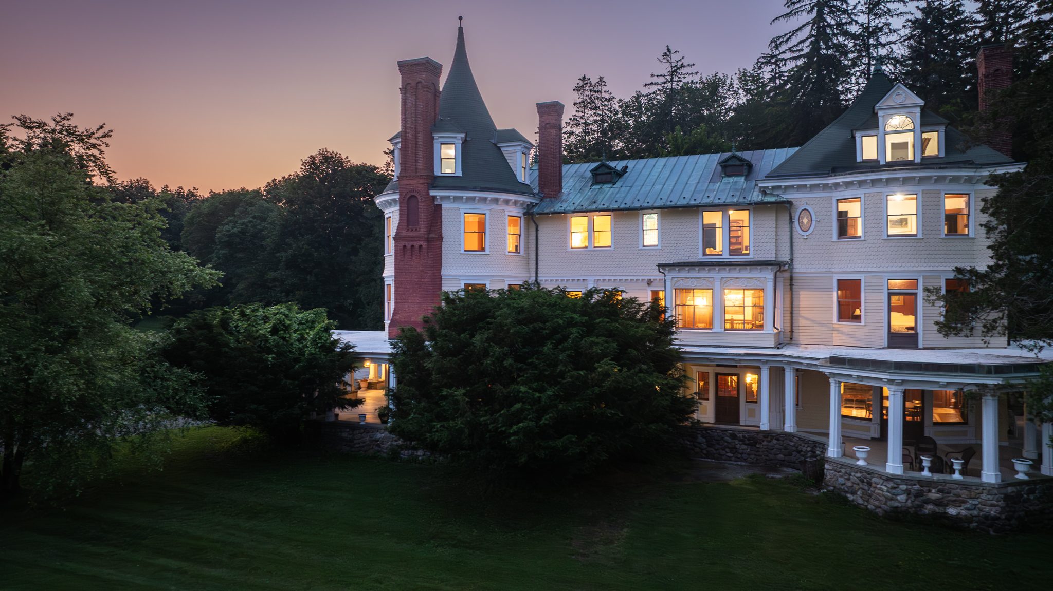 The porch lamps and upstairs windows glowing against a violet sky, the lawn gone almost black in front.