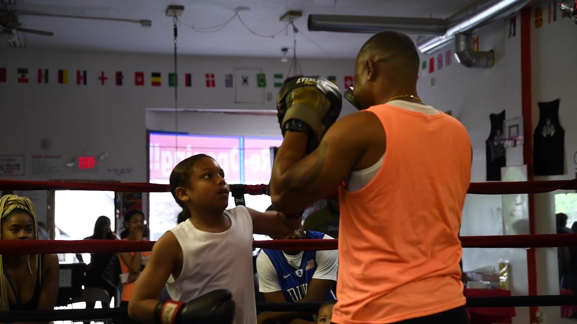 A kid in a white tank drives a straight right into the mitts, spectators watching from the ropes.